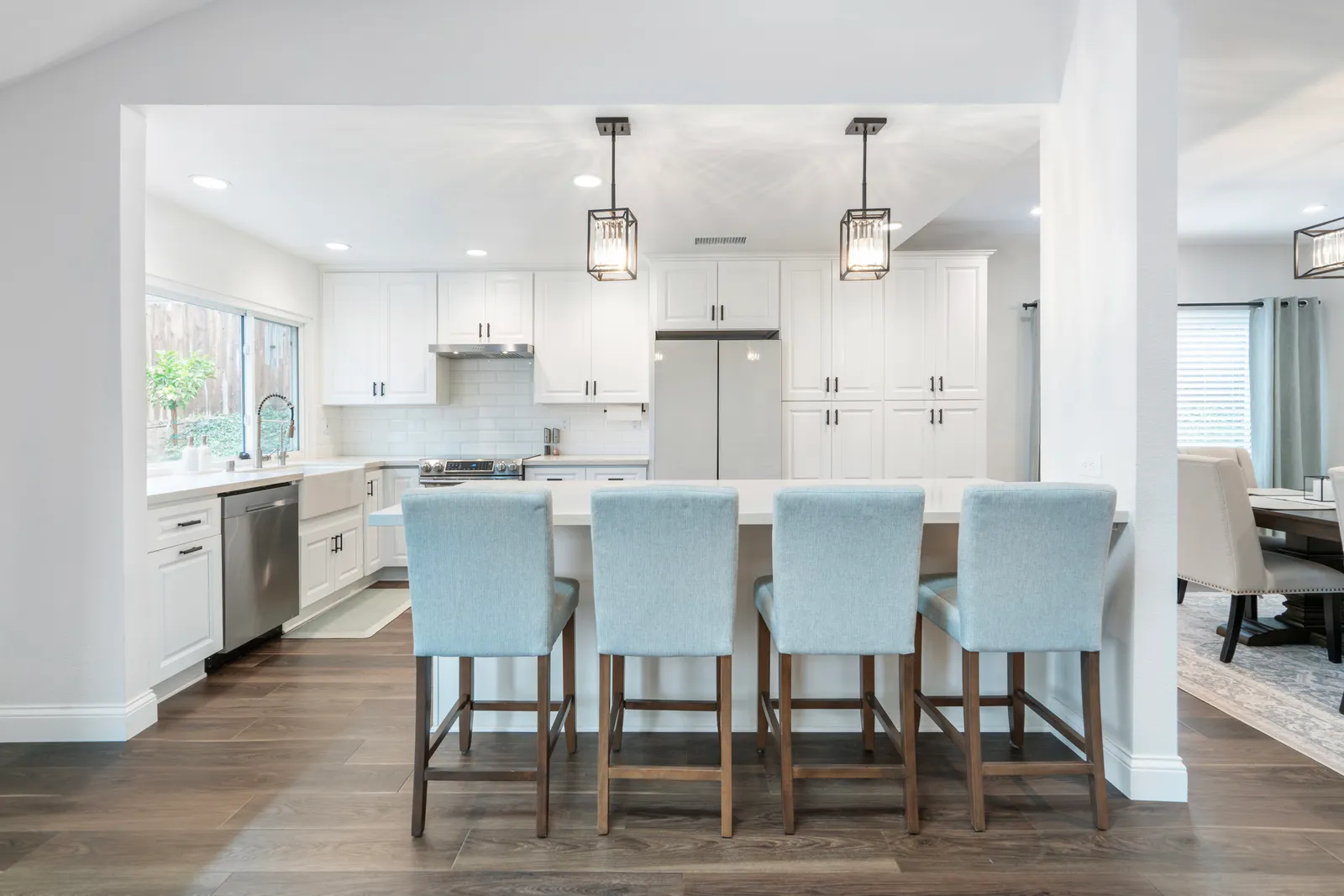 A white kitchen with an island seating four light blue upholstered barstools under black cage pendants.