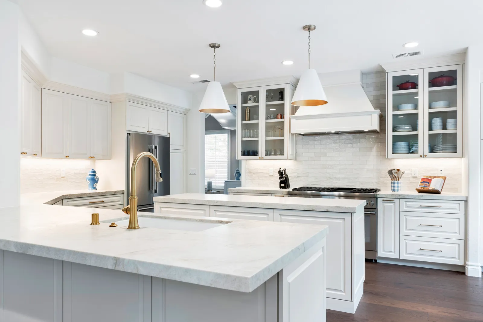 A bright white kitchen with marble countertops, brass faucet, glass-front cabinets, and white cone pendant lights.