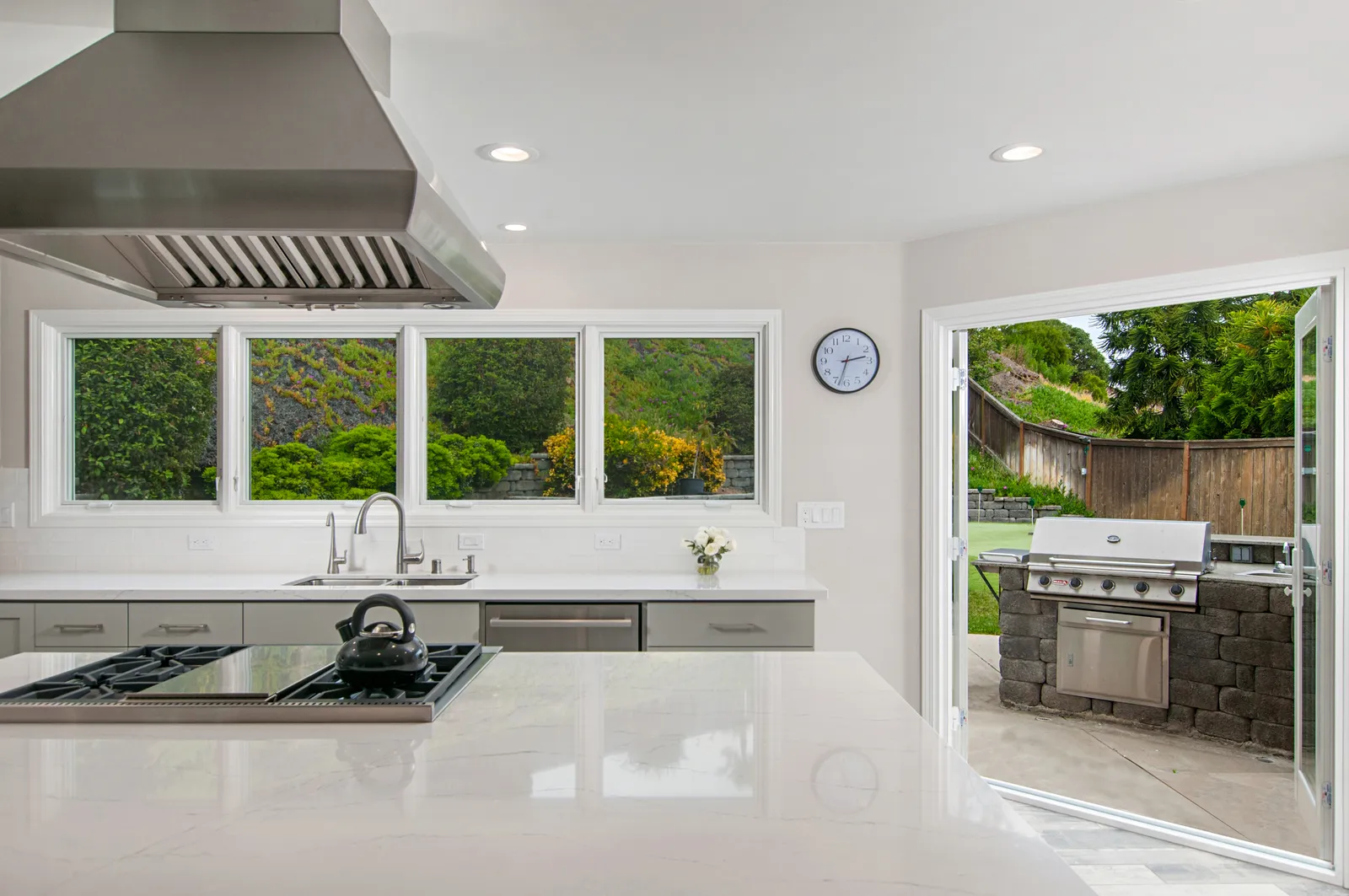 A white kitchen with an island cooktop and stainless hood, opening through French doors to an outdoor grill area.