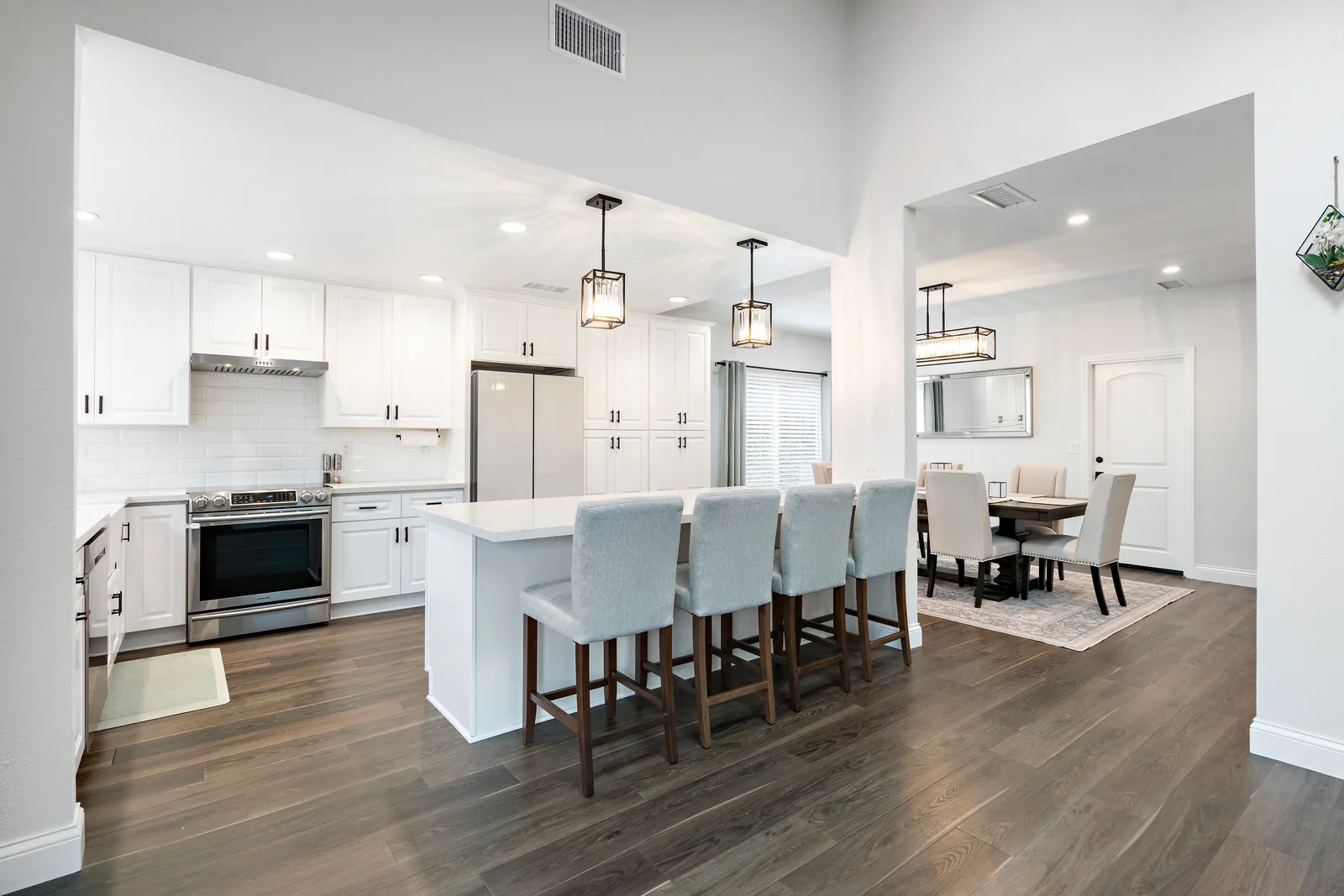 A white kitchen with an island and upholstered bar stools, pendant lights, stainless appliances, and an adjoining dining area.