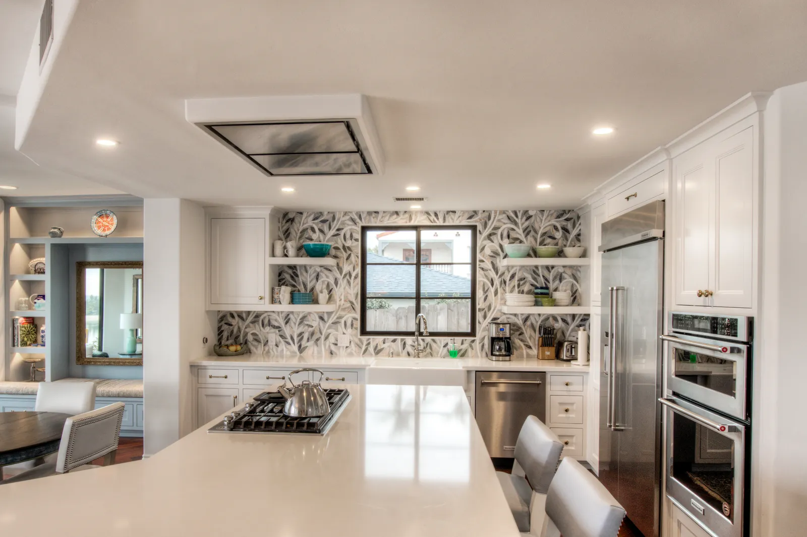A white kitchen with a leaf-patterned mosaic backsplash, farmhouse sink, stainless appliances, and a large island with a gas cooktop.