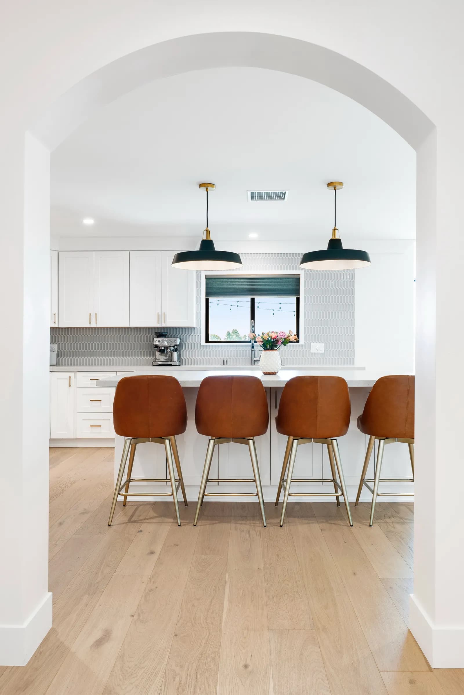 View through an arched opening to a white kitchen island with cognac leather counter stools and dark pendants.