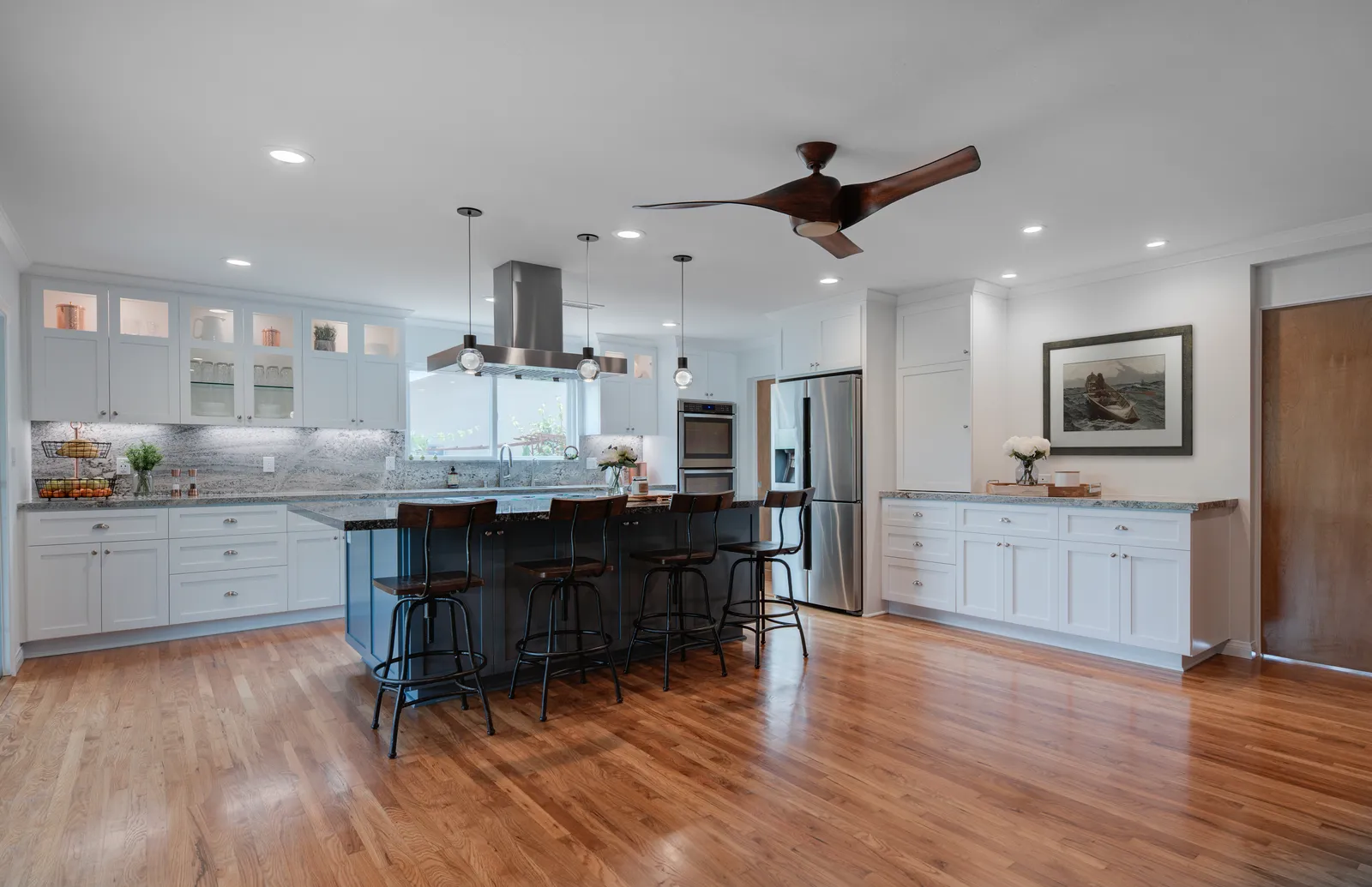 Open white kitchen with glass-front upper cabinets, a navy island with barstools, and oak floors.