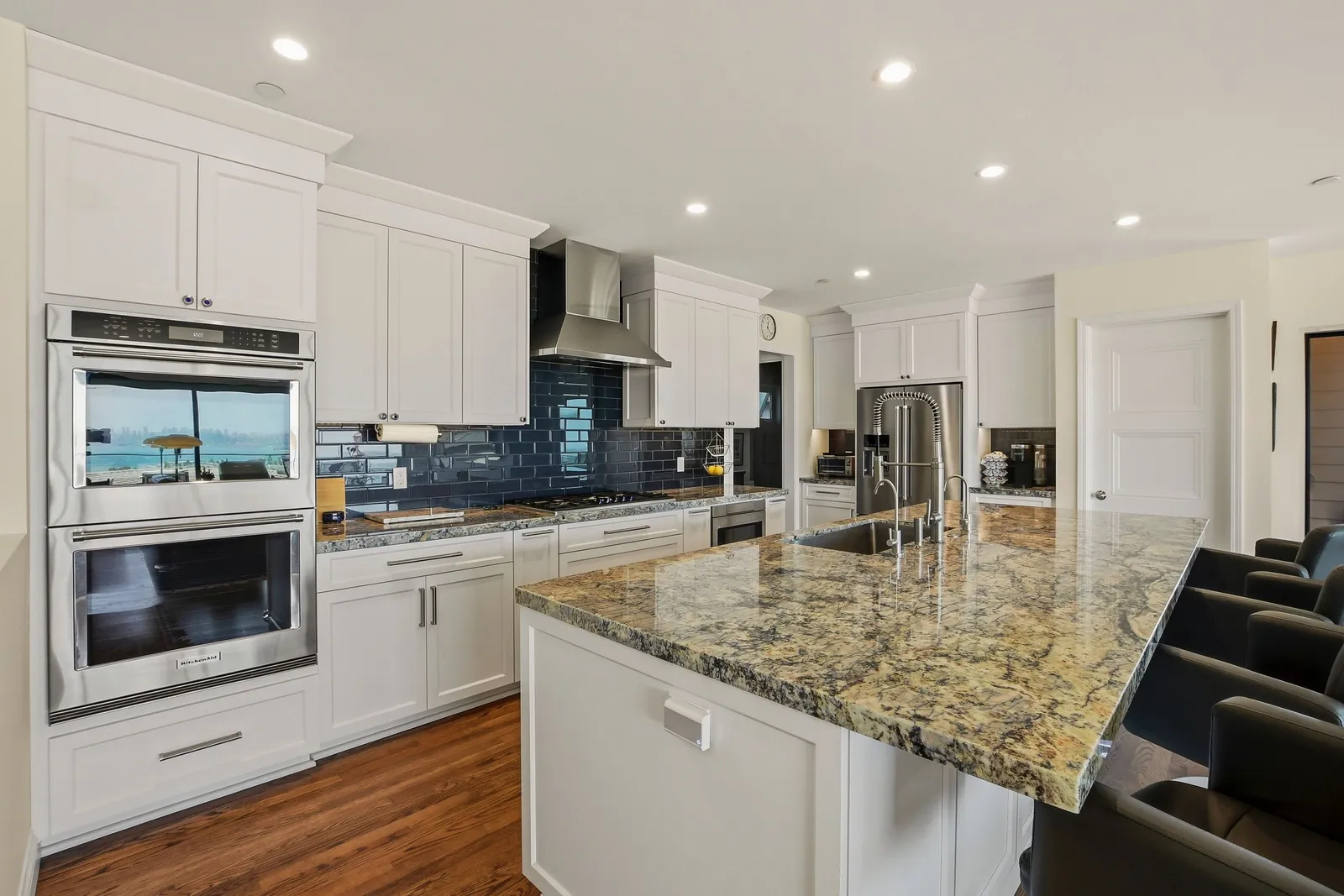 White kitchen with a granite-topped island, glossy navy subway tile backsplash, double wall ovens, and stainless appliances.