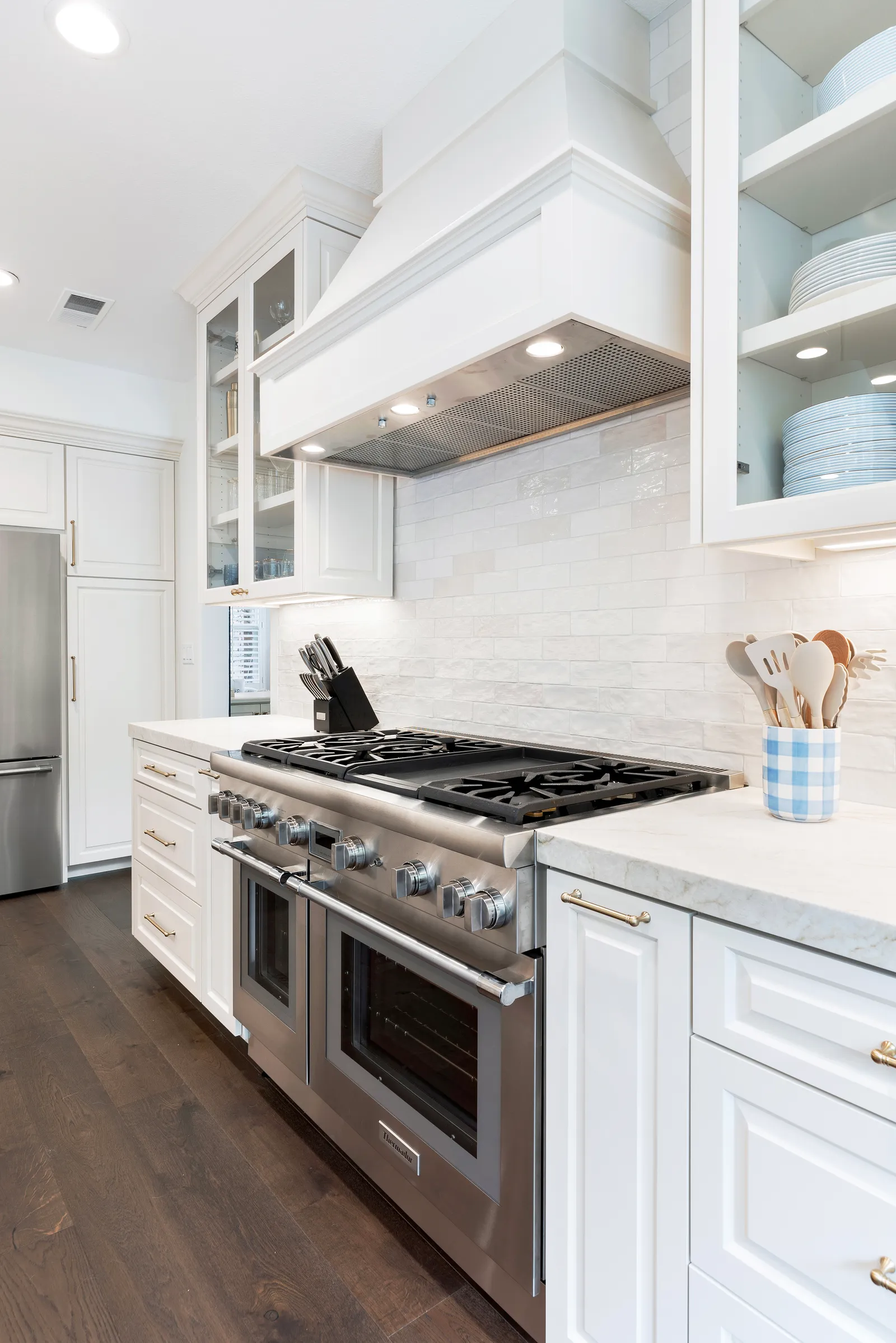 A white kitchen with a stainless professional gas range, custom wood hood, handmade-look subway tile backsplash, and brass hardware.