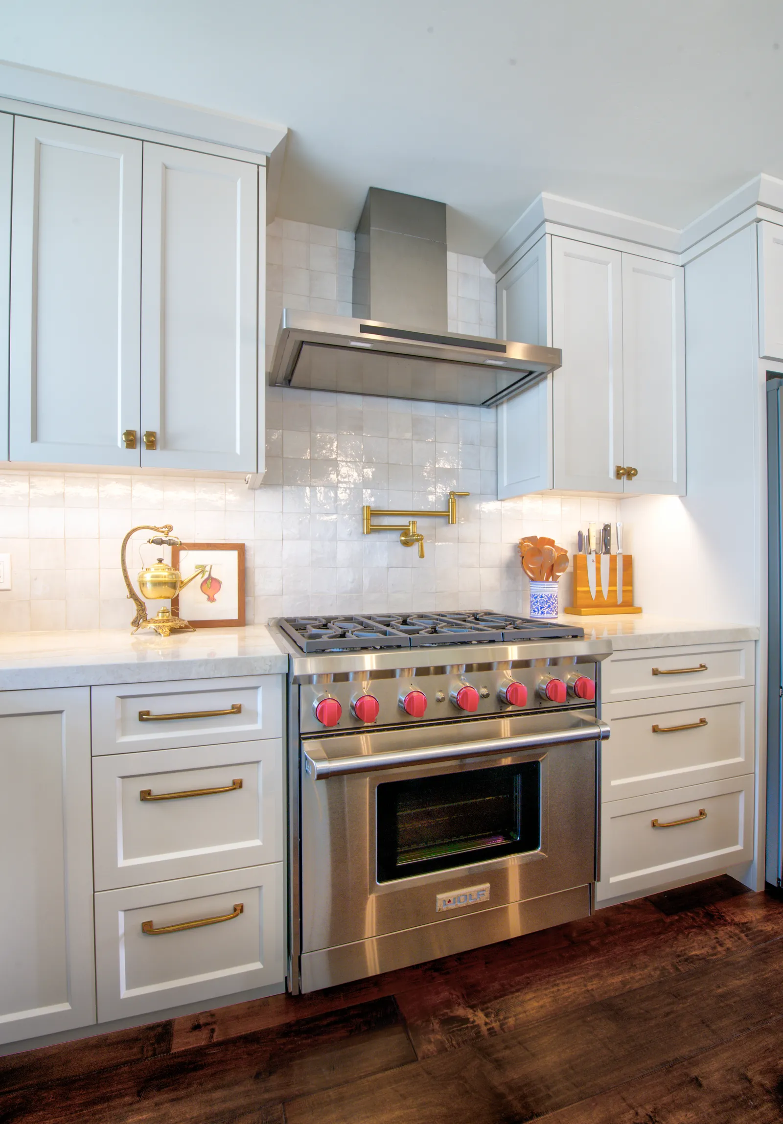 A white shaker kitchen with a stainless professional range, red knobs, brass hardware, and a glossy square tile backsplash.