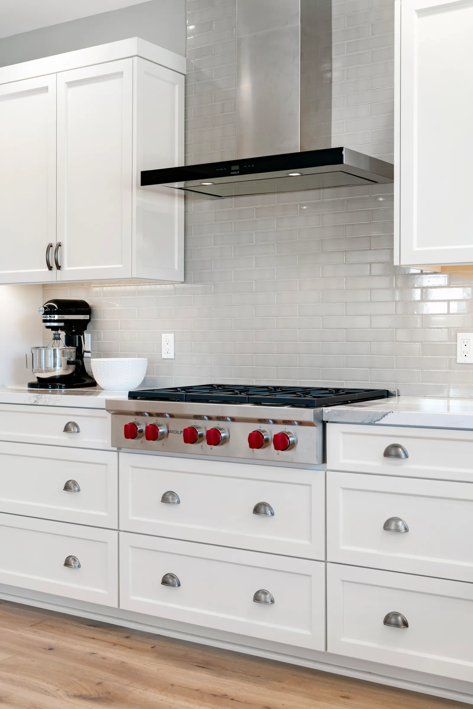White shaker kitchen with cup pulls, a glossy gray subway backsplash, stainless chimney hood, and a professional rangetop with red knobs.