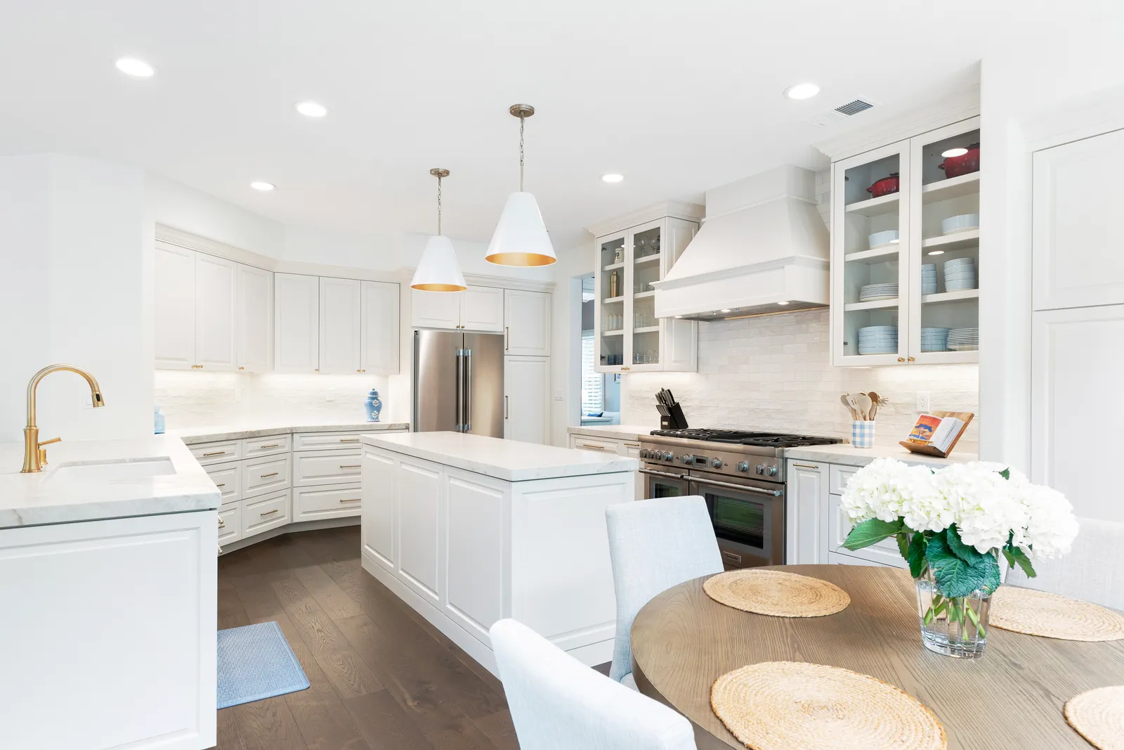 A white kitchen with marble island, brass faucet, professional range, and a round wood breakfast table with hydrangeas.