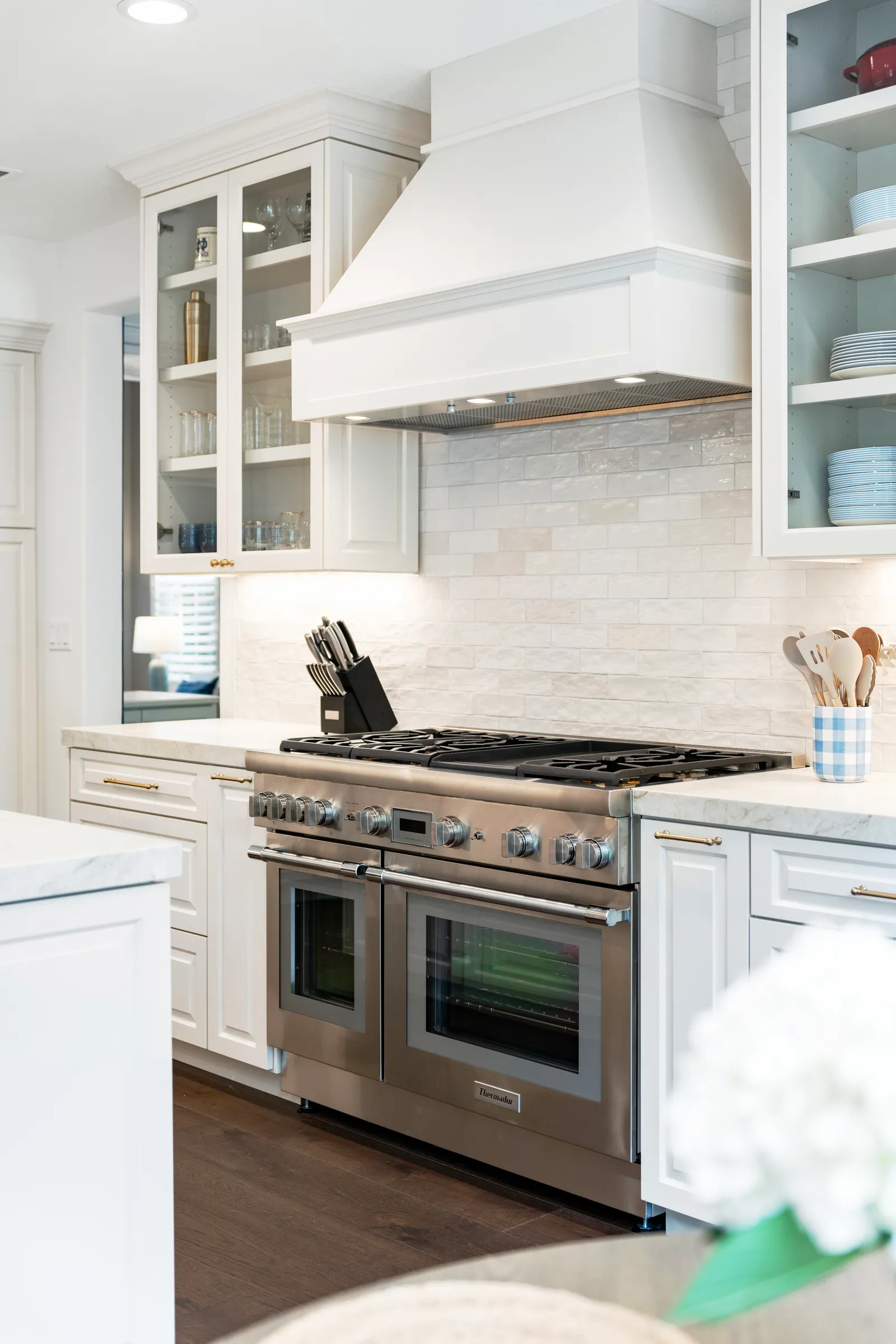 A white kitchen with a shaker-style range hood, glossy subway tile, glass-front cabinets, and a stainless double-oven range.