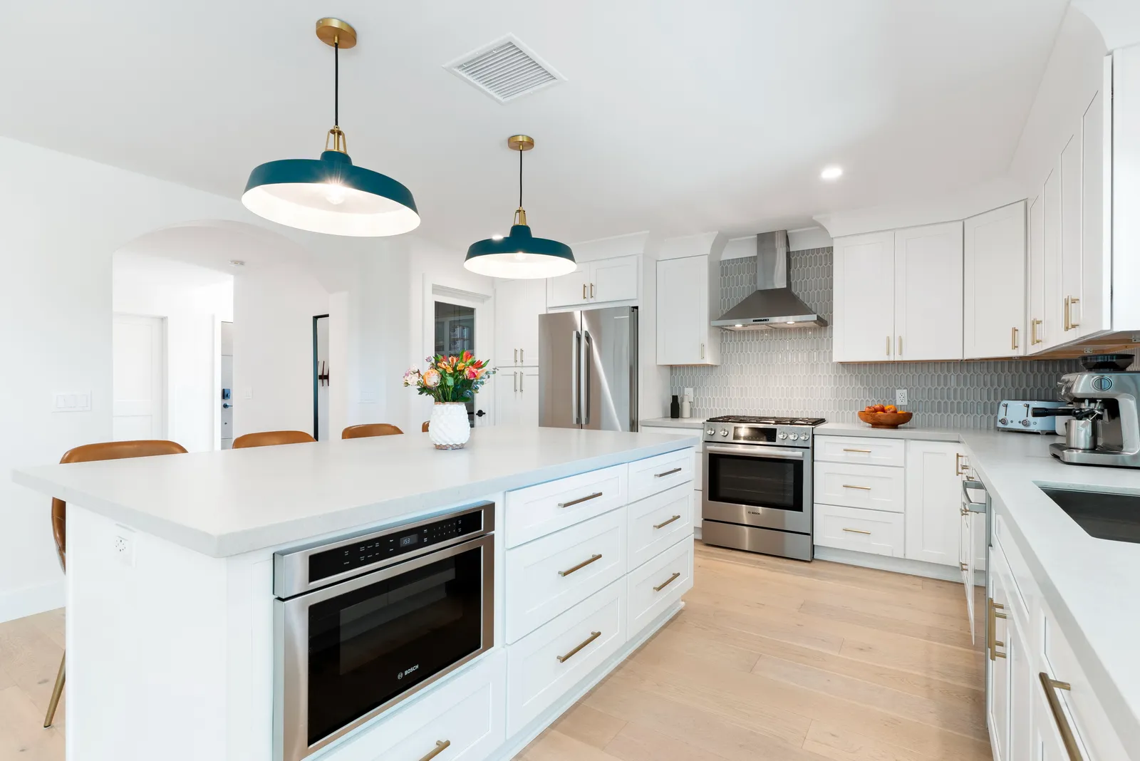 White shaker kitchen with a large island, microwave drawer, teal dome pendants, and light oak flooring.