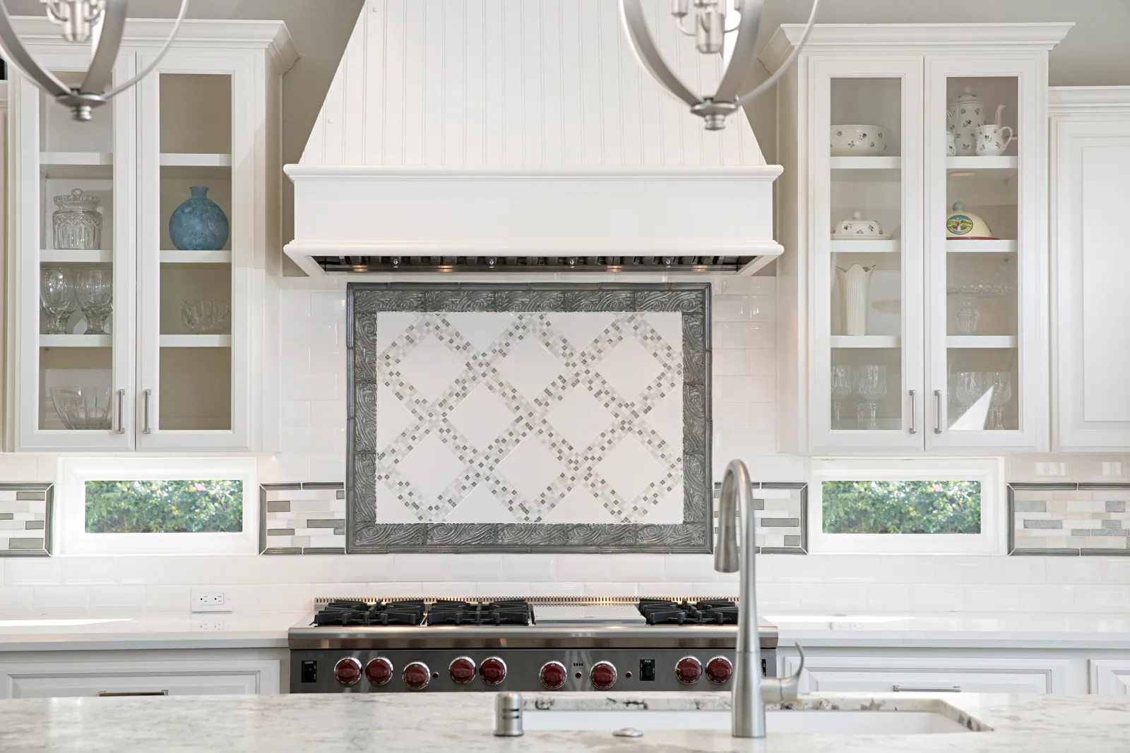 A white kitchen with a beadboard range hood, mosaic tile medallion backsplash, glass-front cabinets, and a professional range.