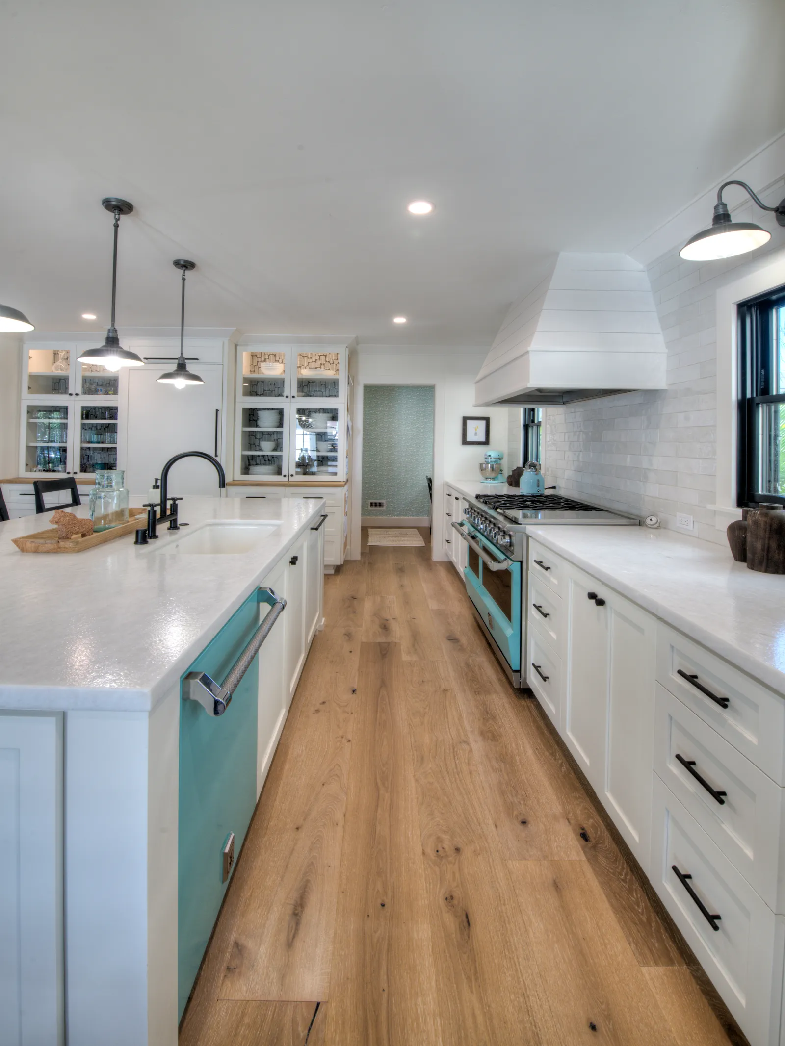 A bright white kitchen with a large island, glass-front cabinets, wide-plank oak floors, and a retro turquoise range and dishwasher.