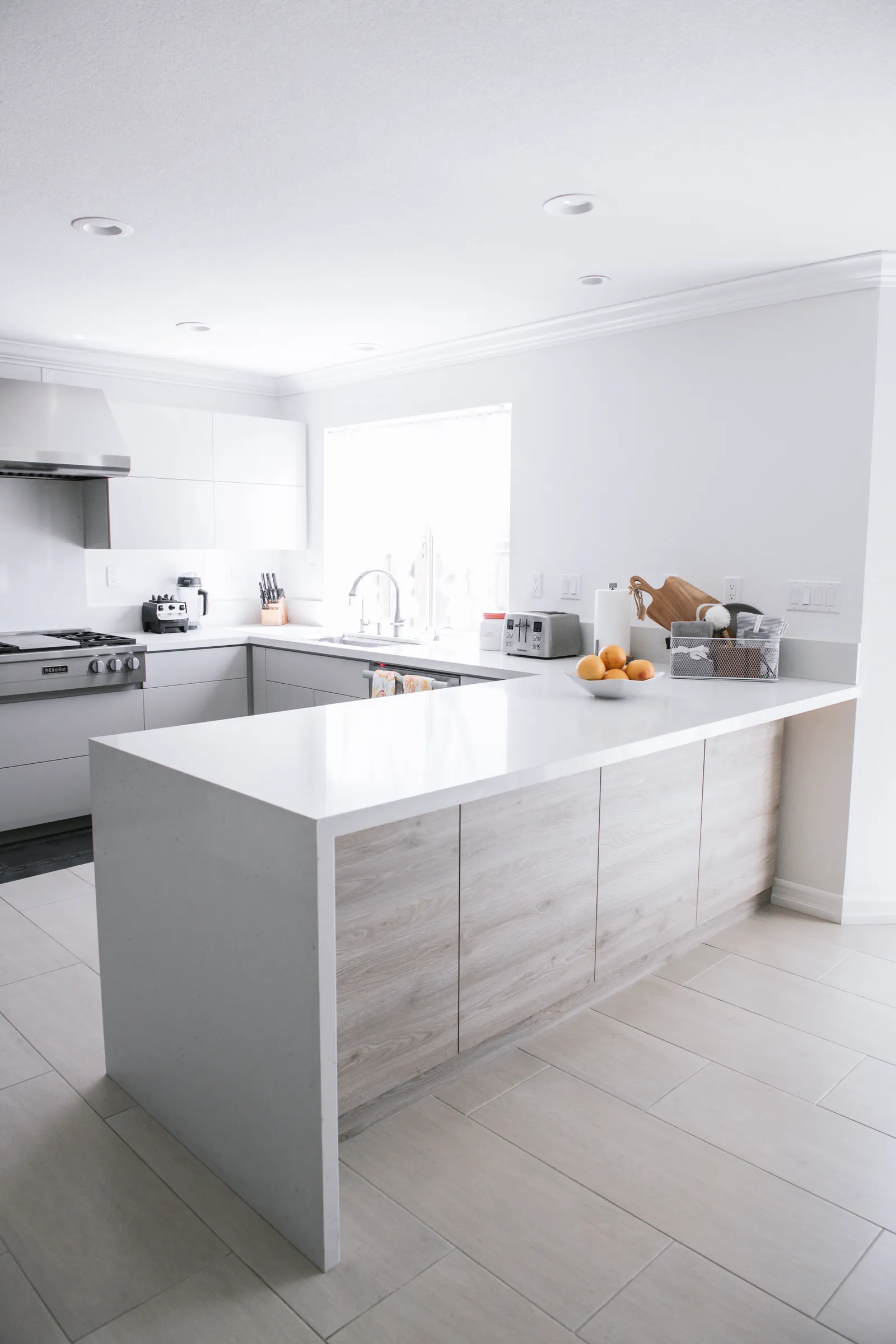 Bright minimalist kitchen with white waterfall-edge peninsula, light wood cabinet fronts, and a professional range.