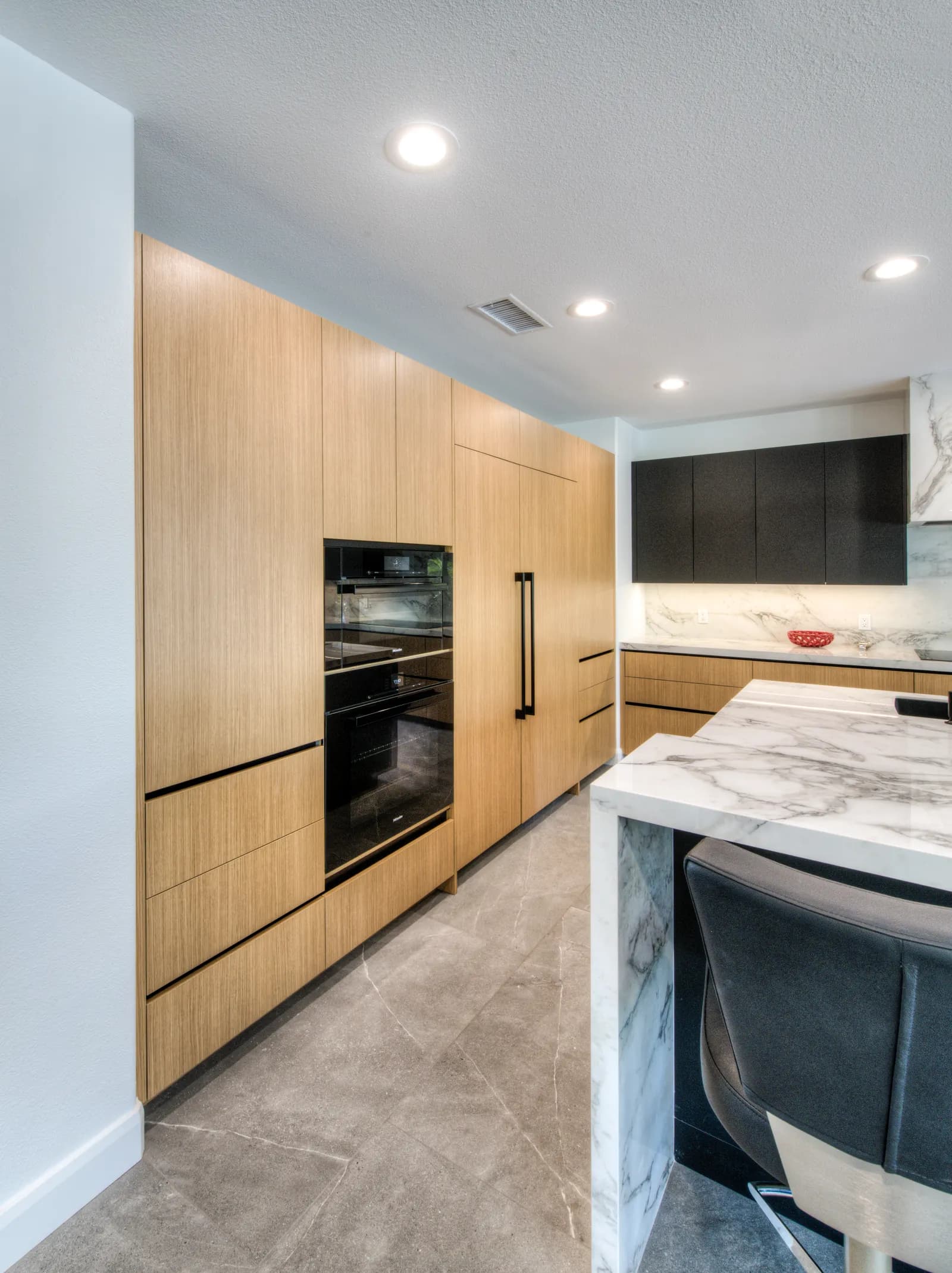Modern kitchen with flat-panel white oak cabinets, black accents, and a marble waterfall island with a leather stool.
