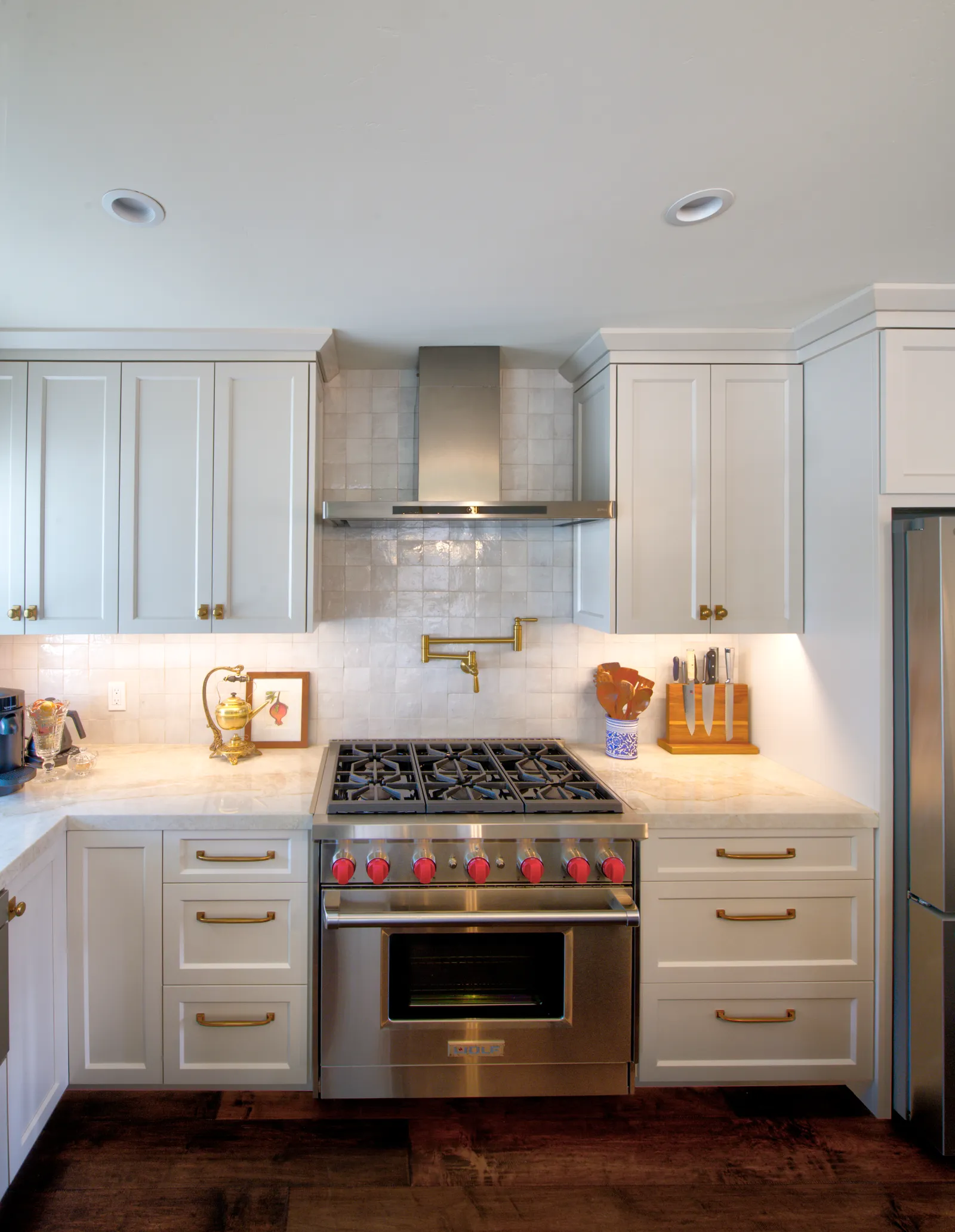 A white shaker kitchen with brass hardware, glossy square tile backsplash and a professional gas range with red knobs.