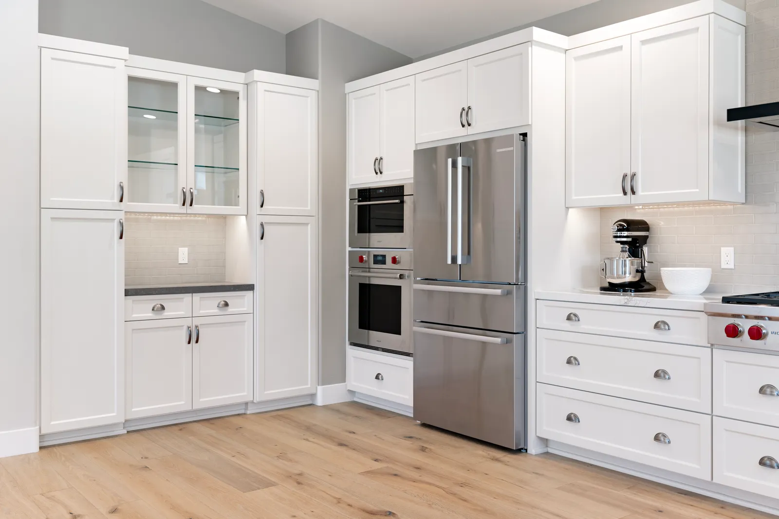 A white shaker kitchen with stacked wall ovens, a counter-depth stainless refrigerator, and light oak floors.