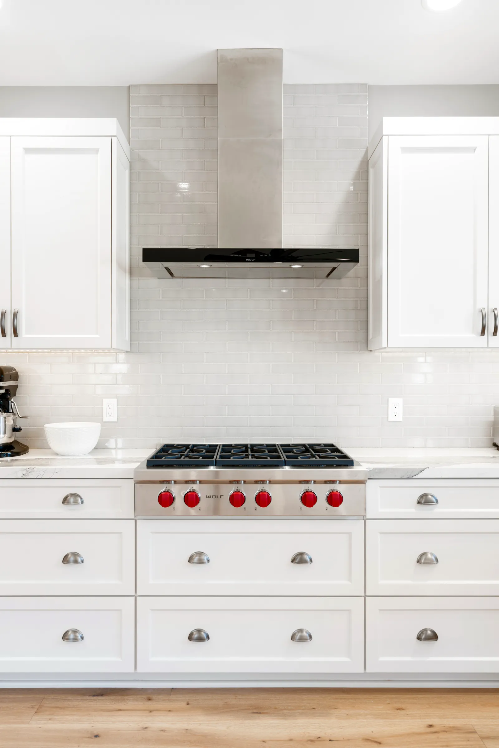 A white kitchen with shaker cabinets, subway tile backsplash, stainless hood, and a pro-style gas rangetop with red knobs.