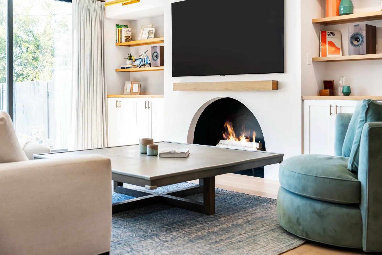 Living room with an arched fireplace below a wall-mounted TV, oak mantel shelf, built-in shelving, and a blue swivel chair.