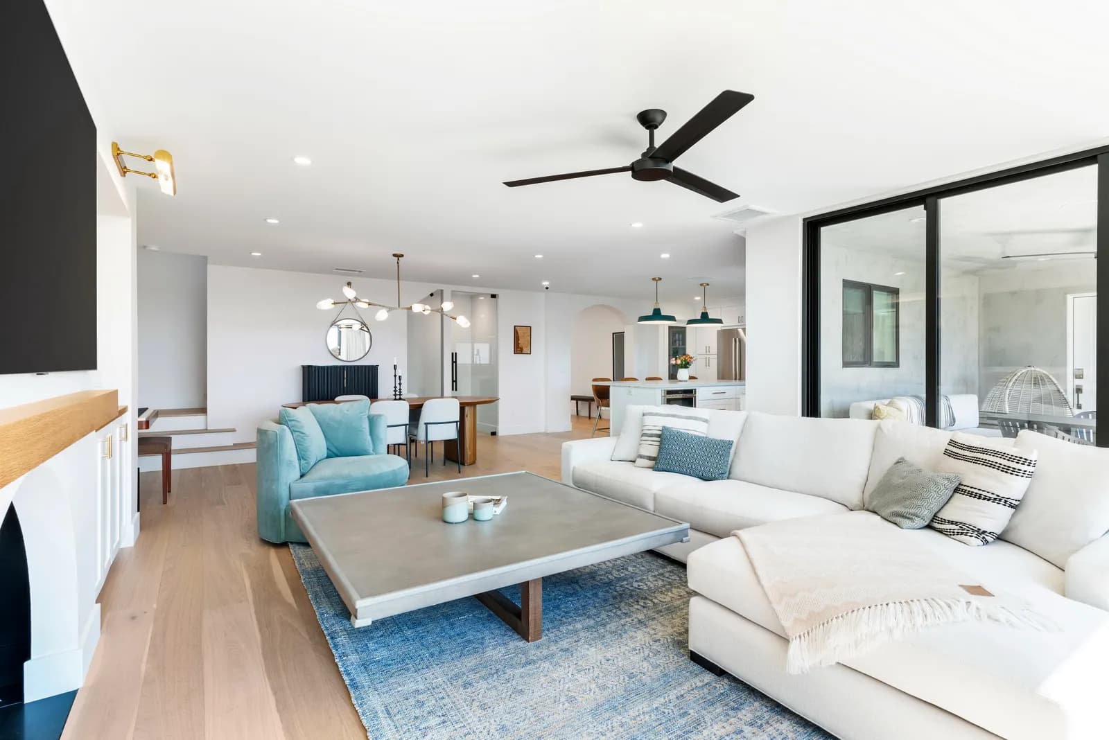 A bright open-plan living room with a white sectional sofa, concrete coffee table, blue rug, and sliding glass doors.