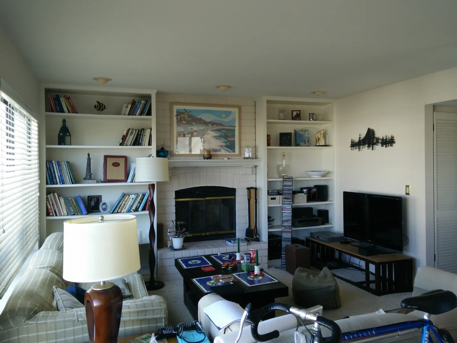 A lived-in family room with white built-in bookshelves, a painted brick fireplace, a wall-mounted TV, and a bicycle in the foreground.