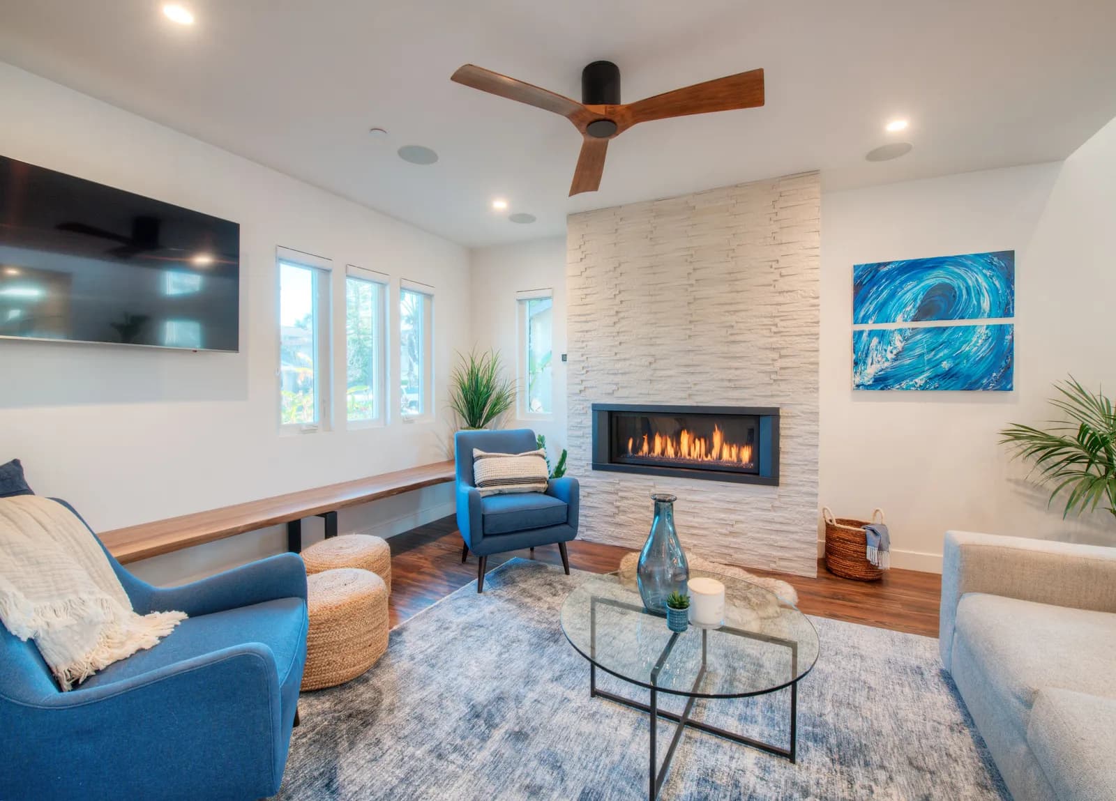 A coastal-styled living room with a white stacked-stone fireplace wall, linear gas fireplace, blue armchairs, and a wood ceiling fan.
