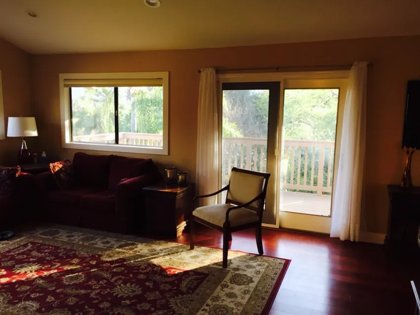 A dated living room with a burgundy sofa, oriental rug, and a sliding door opening to a wood deck railing.