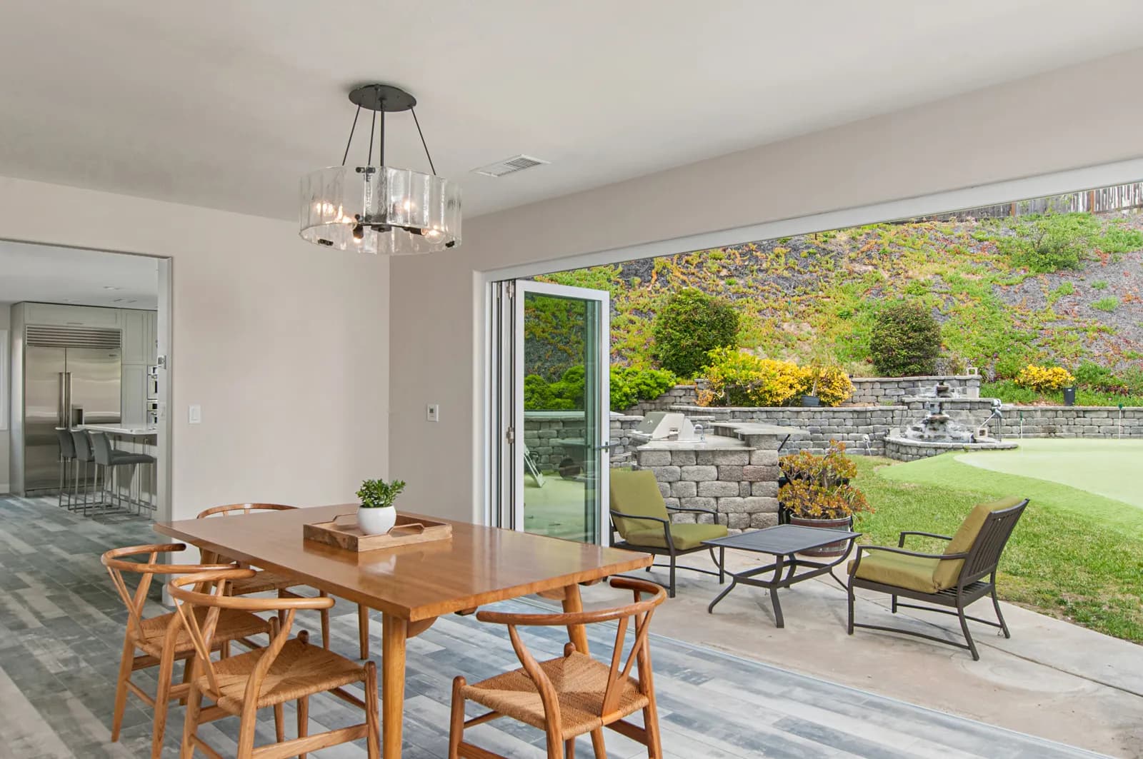 Dining area with wishbone chairs opening through folding glass doors to a terraced backyard patio.