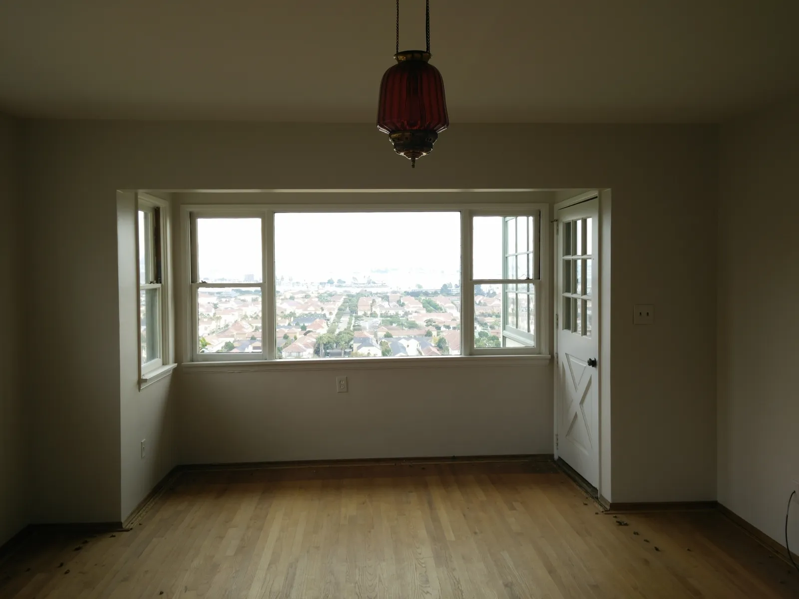 An empty room with oak floors and a large picture window overlooking rooftops, with a vintage red glass pendant light hanging from the ceiling.
