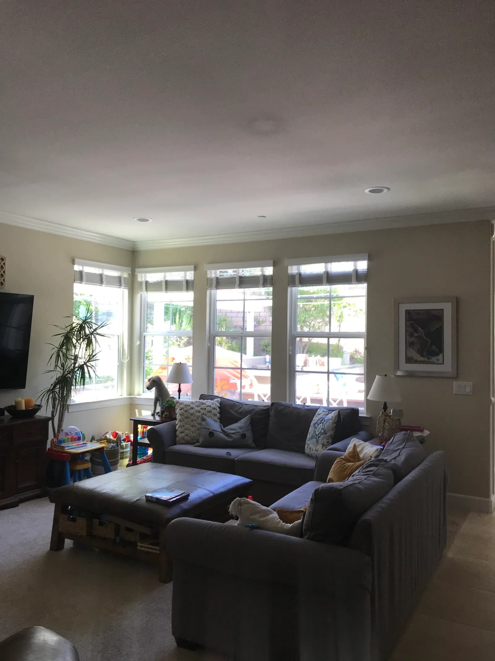 A family room with gray sofas, a leather ottoman coffee table, corner windows, and children's toys along the wall.