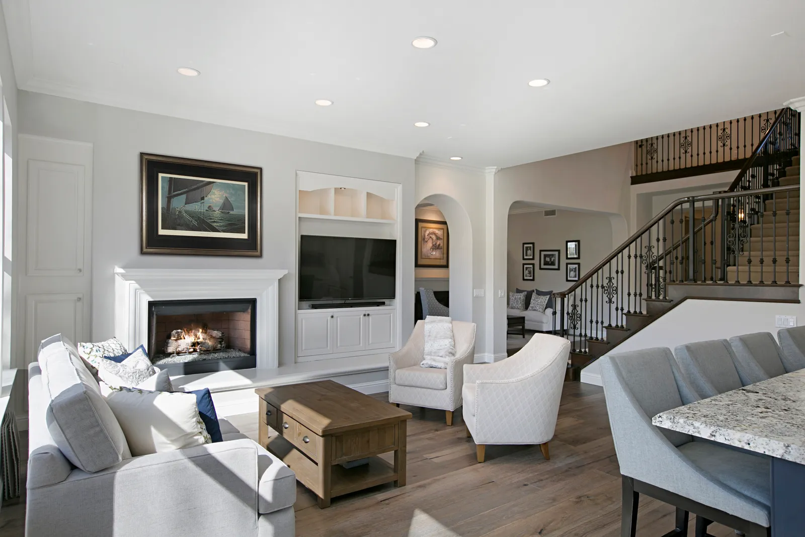 Living room with white mantel fireplace, built-in media cabinet, neutral armchairs, and a wrought iron staircase beyond.