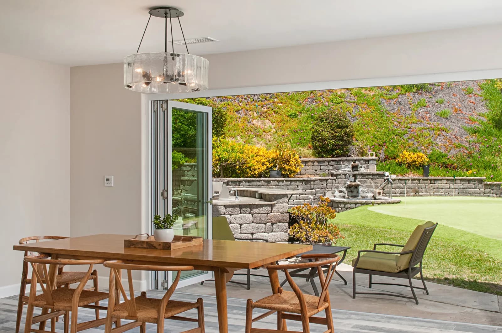Dining area with wishbone chairs opening through folding glass doors to a backyard putting green.