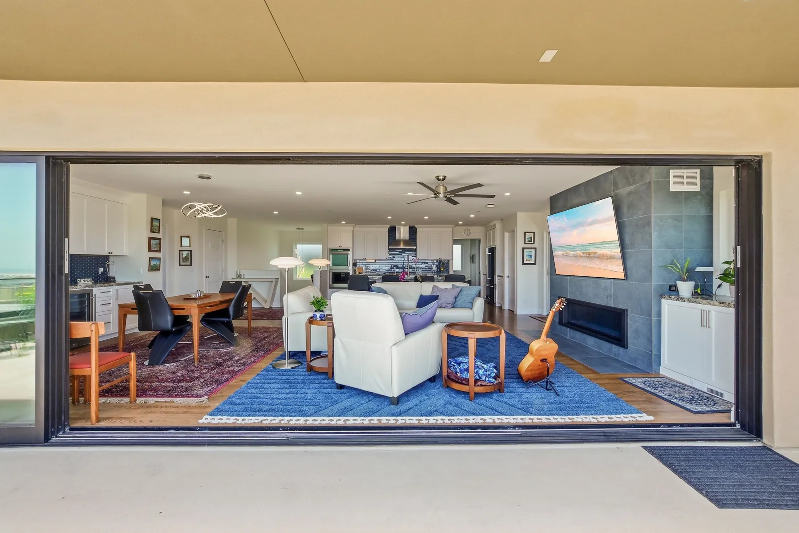 View through a fully opened glass wall into a great room with white leather seating, blue rug, dining area, and slate fireplace wall.