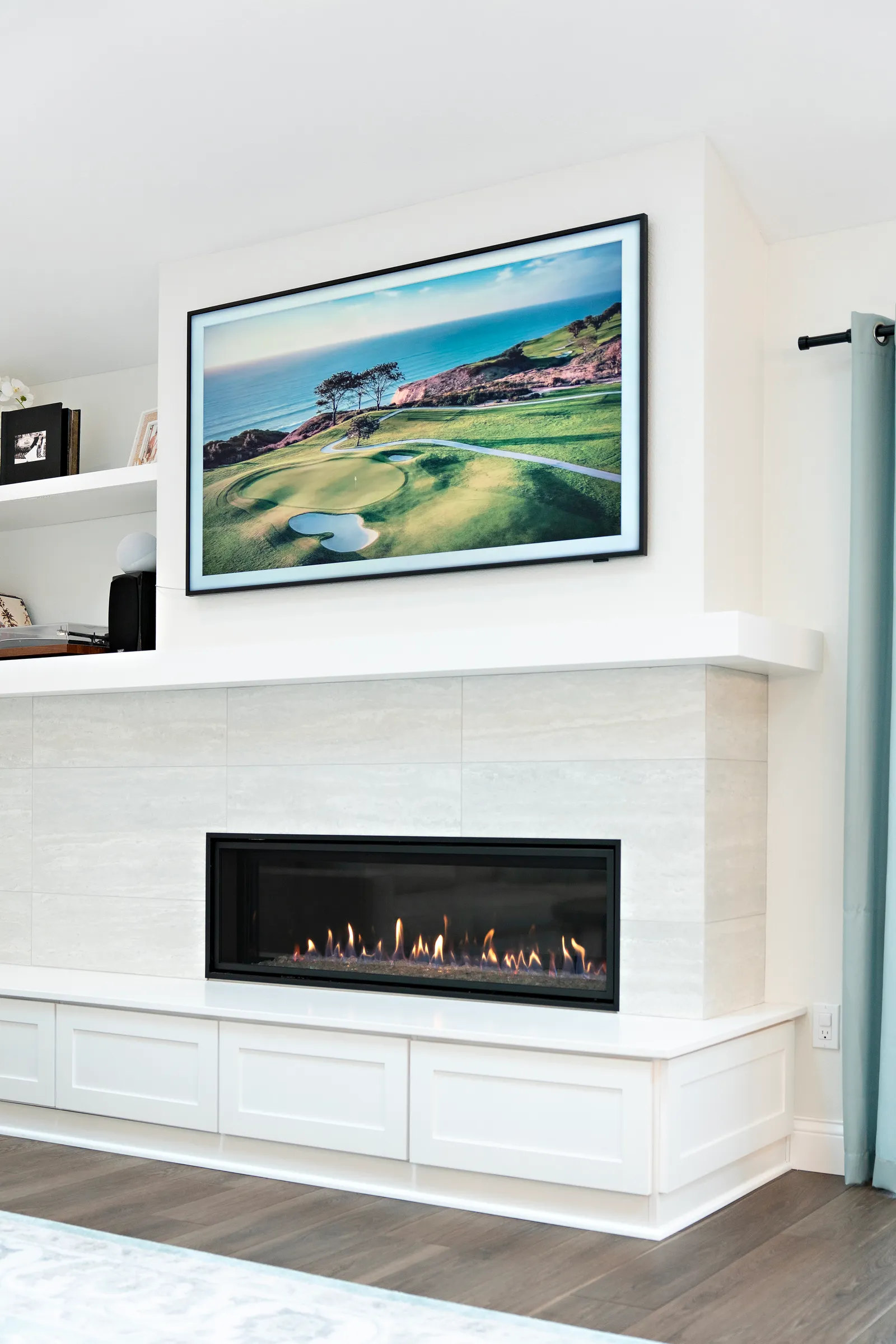 A modern media wall with a linear gas fireplace, light stone-look tile, floating white shelves, and a framed television above.