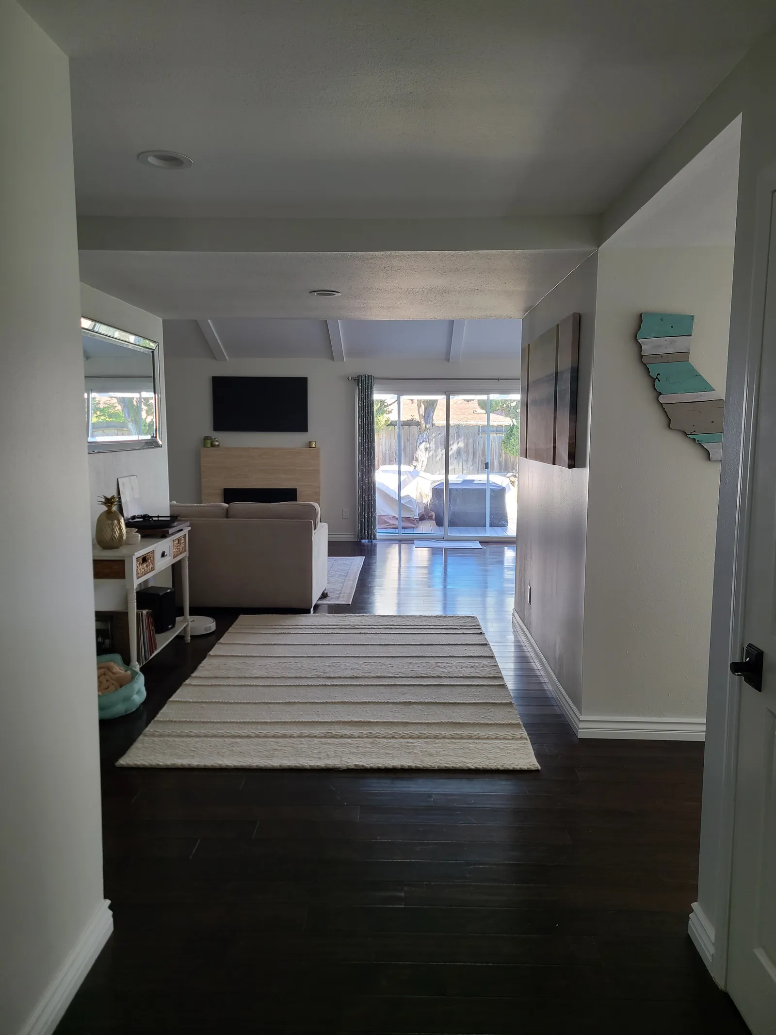 A living room with dark wood floors, a striped rug, beige sofa and a sliding glass door to a fenced backyard.