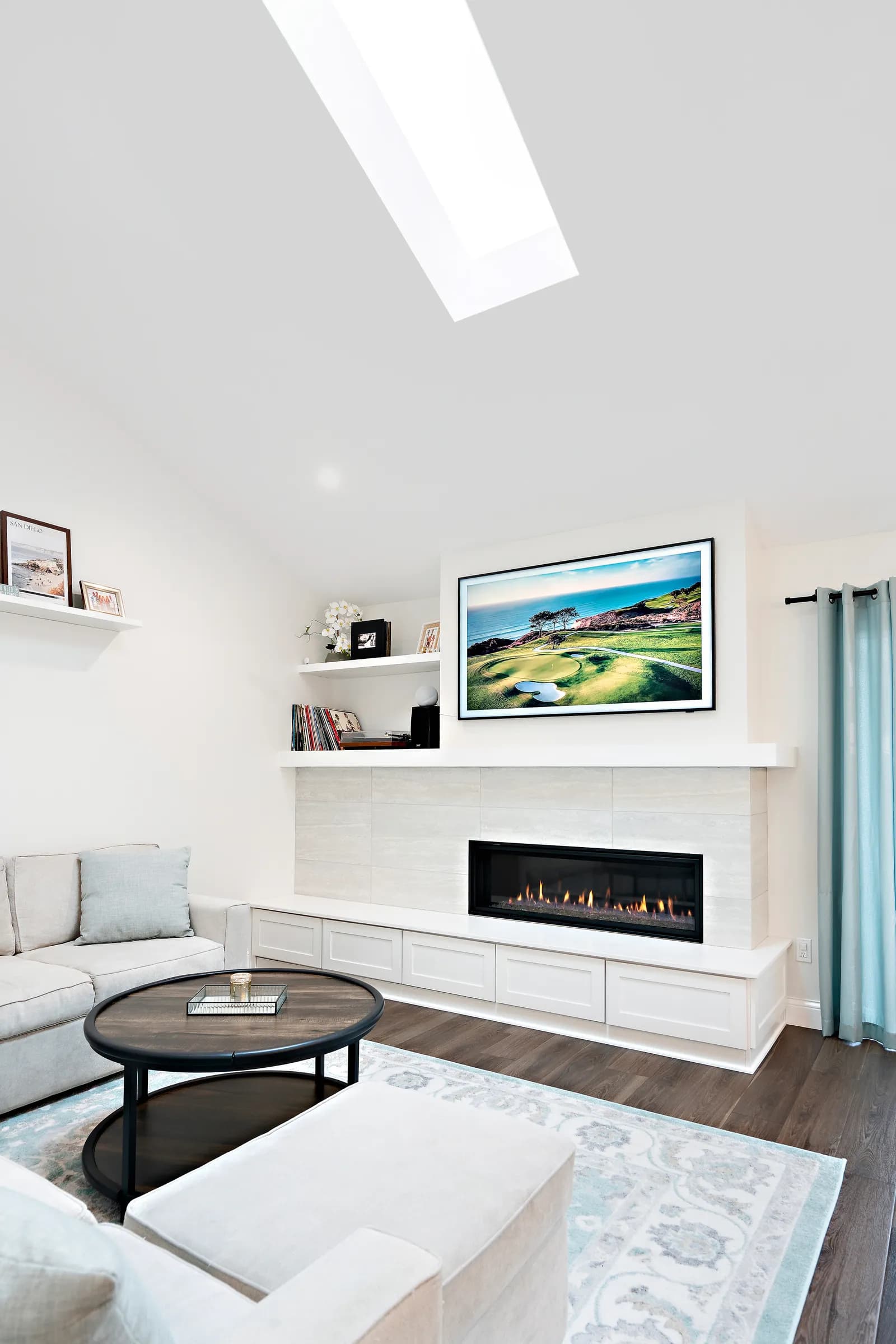 Living room with a linear gas fireplace, tile surround, wall-mounted TV, skylight, and white built-in bench cabinets.