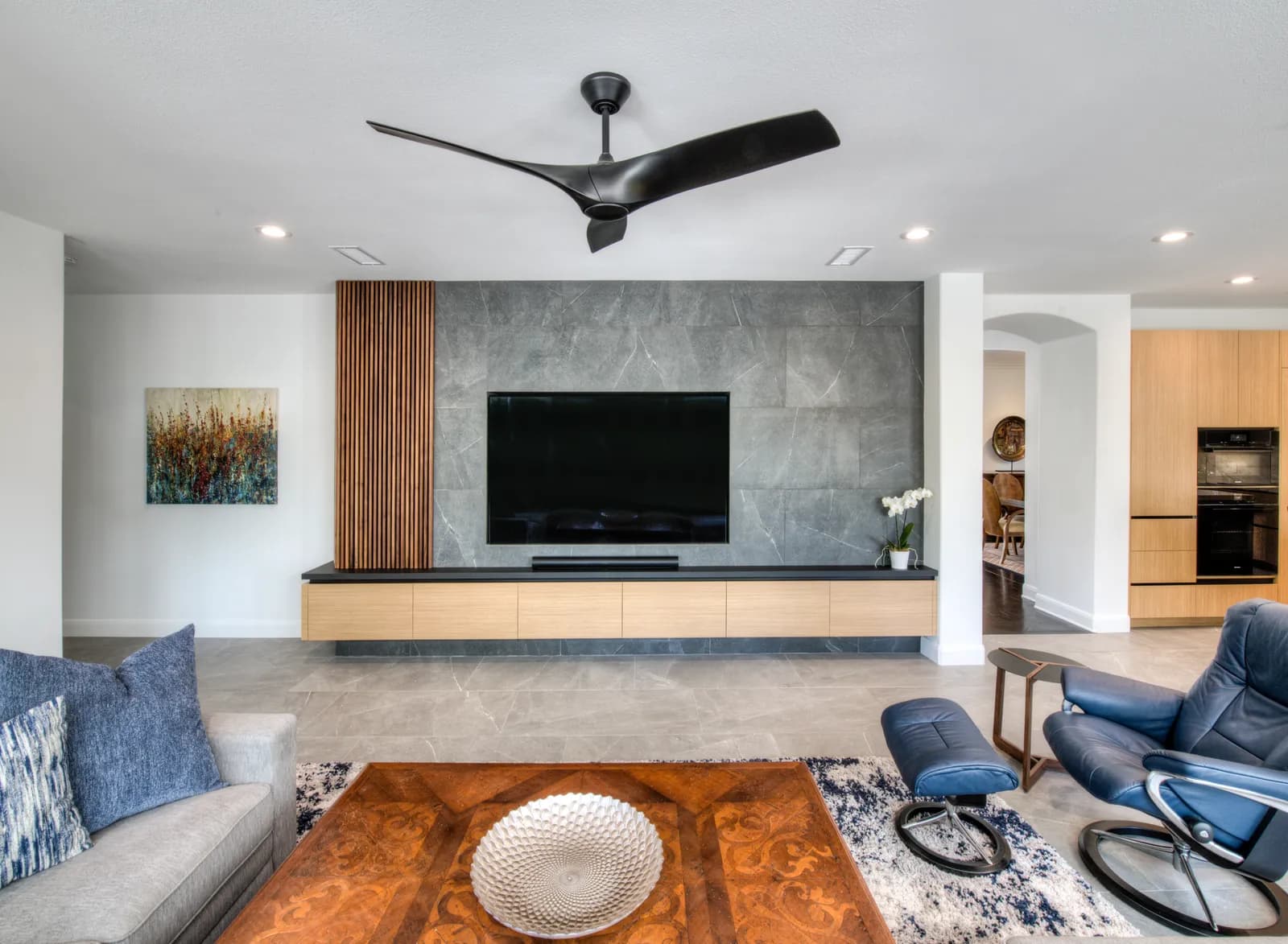 Modern living room with gray stone media wall, wood slat accent, floating oak console, and blue leather recliners.