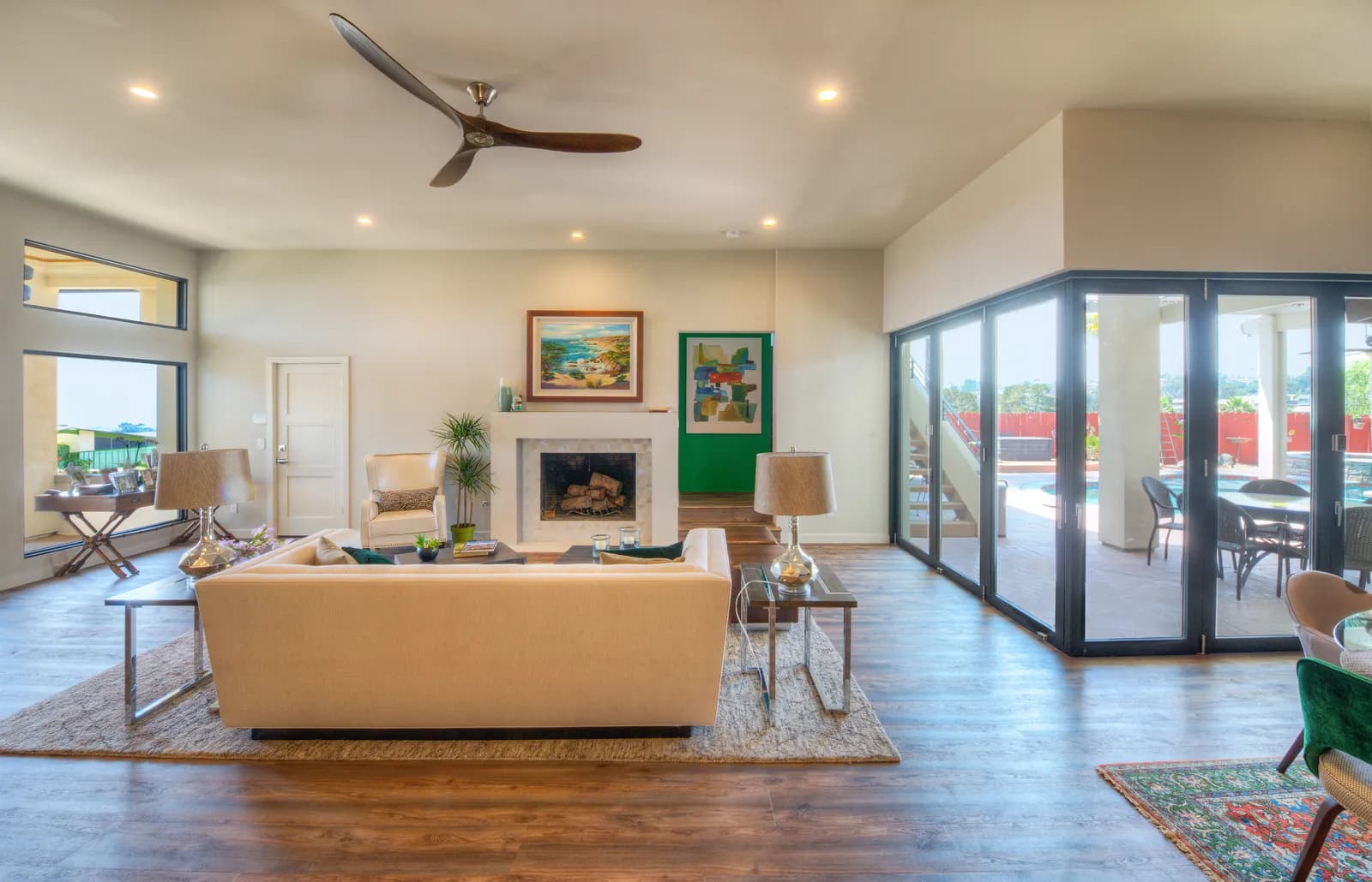 An open living room with a ceiling fan, fireplace, and black-framed bifold glass doors opening to a pool patio.
