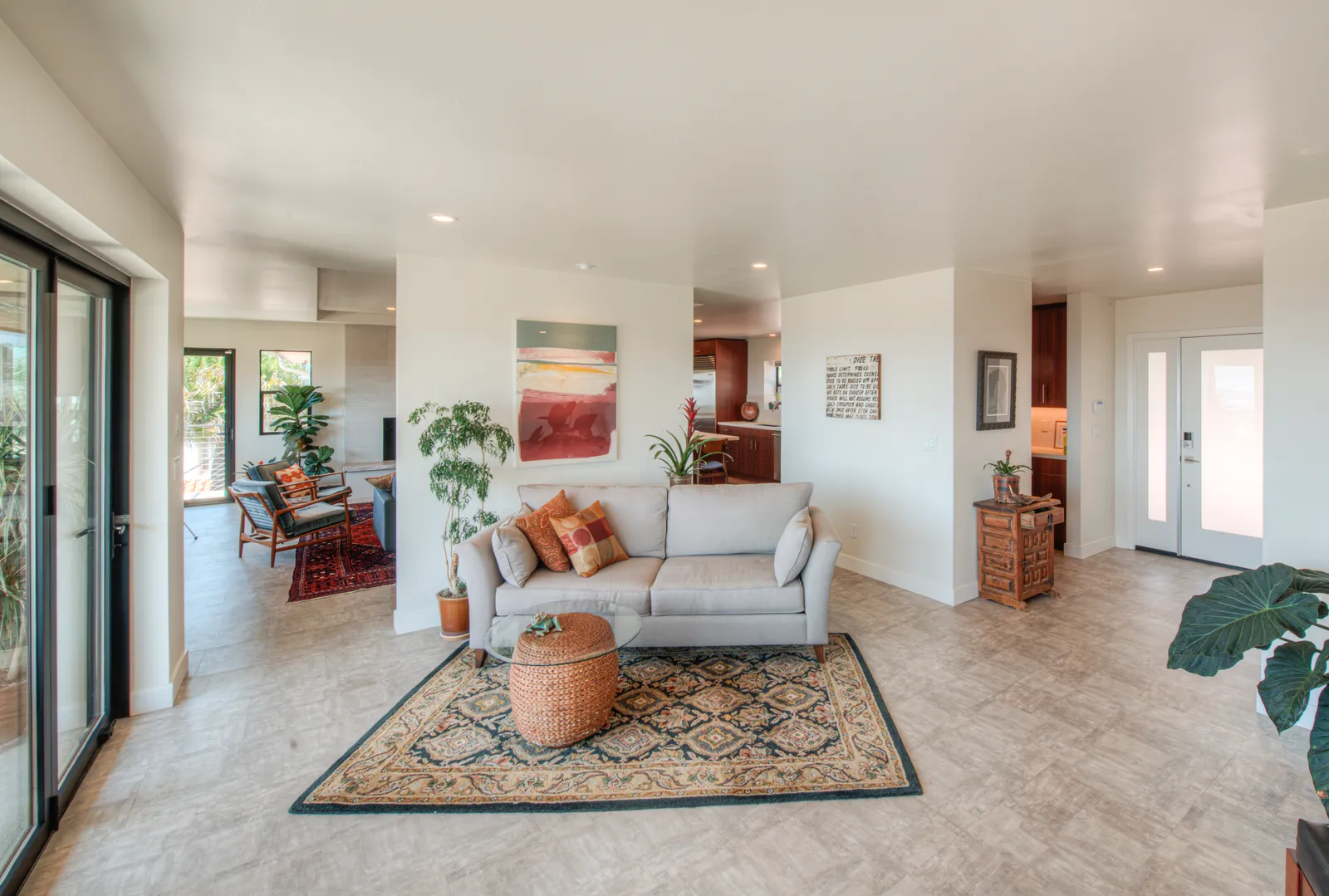 Open-plan living room with a light gray sofa, patterned area rug, potted plants, and sliding glass doors to a patio.