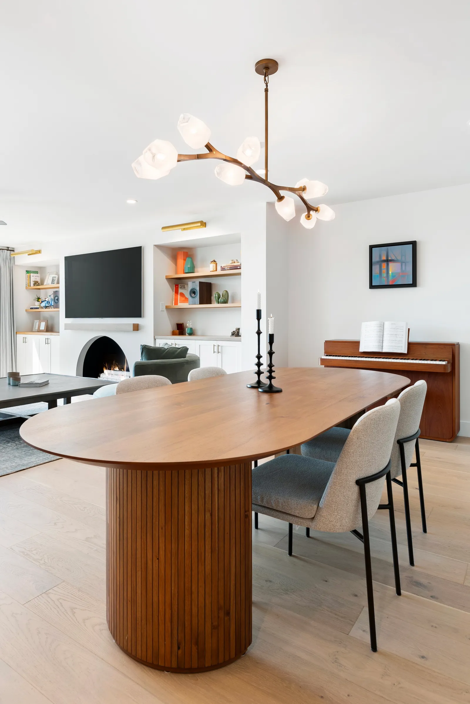 A dining area with an oval walnut table on a fluted pedestal base, upholstered chairs, a branch chandelier, piano, and arched fireplace.