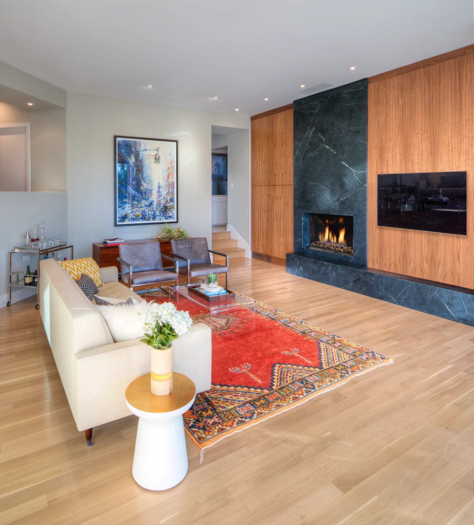 Modern living room with a soapstone fireplace surround, walnut paneled TV wall, and a red patterned rug.