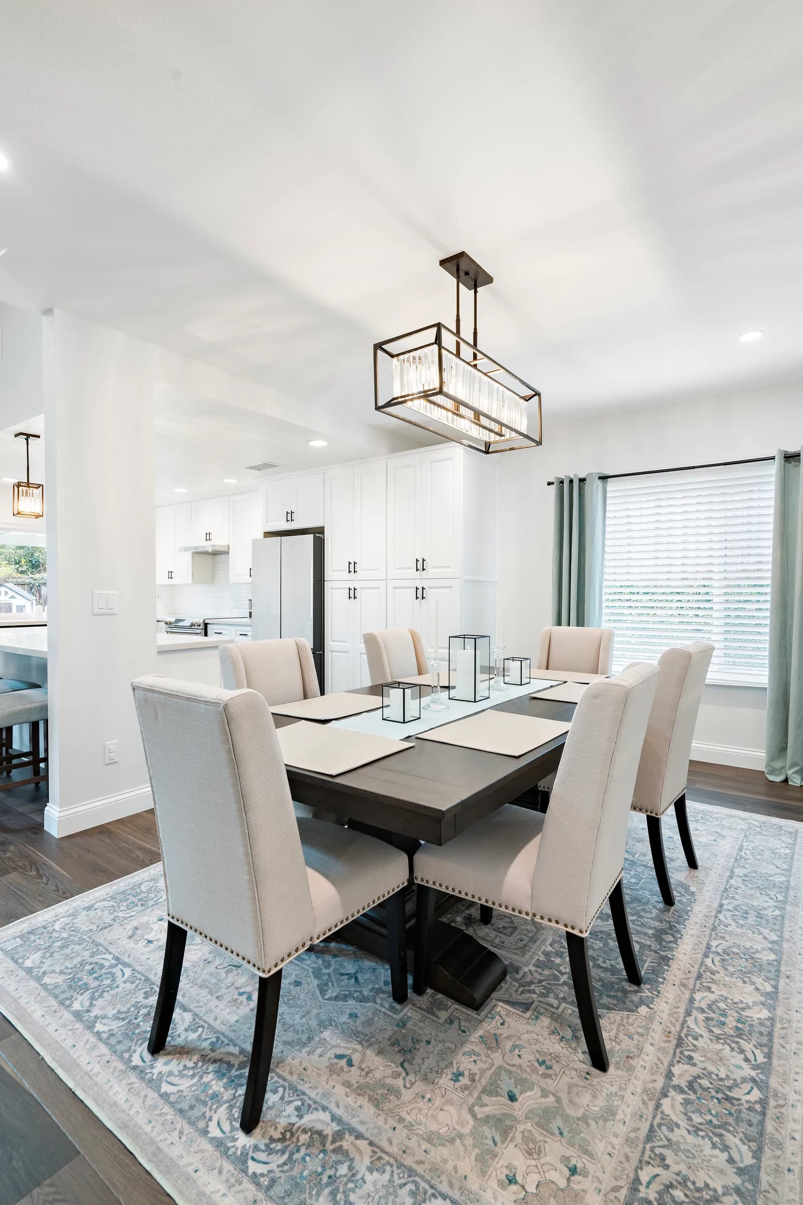 A staged dining room with upholstered chairs, a dark wood table with candle centerpieces, and a rectangular crystal chandelier.