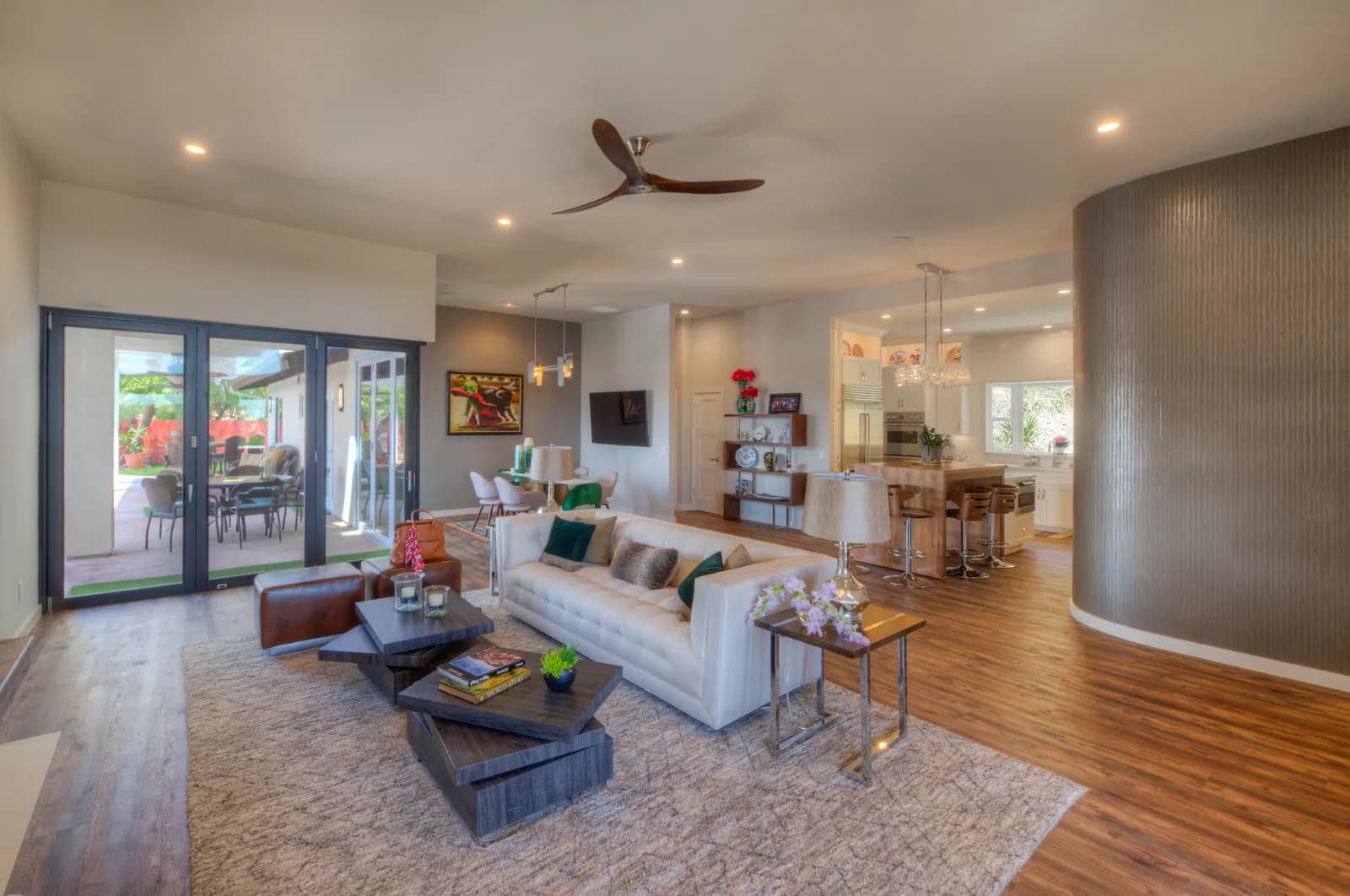 A staged open-plan living room with a white tufted sofa, layered rug, ceiling fan, and folding glass doors to a patio.