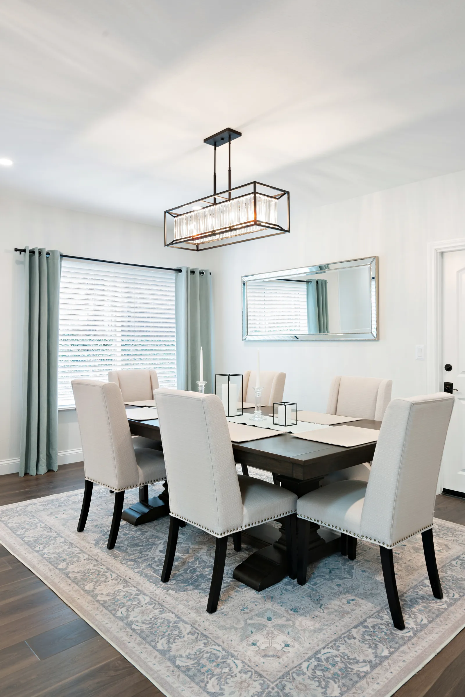 Staged dining room with an upholstered chair set, dark wood table, rectangular crystal chandelier, and a large beveled mirror.