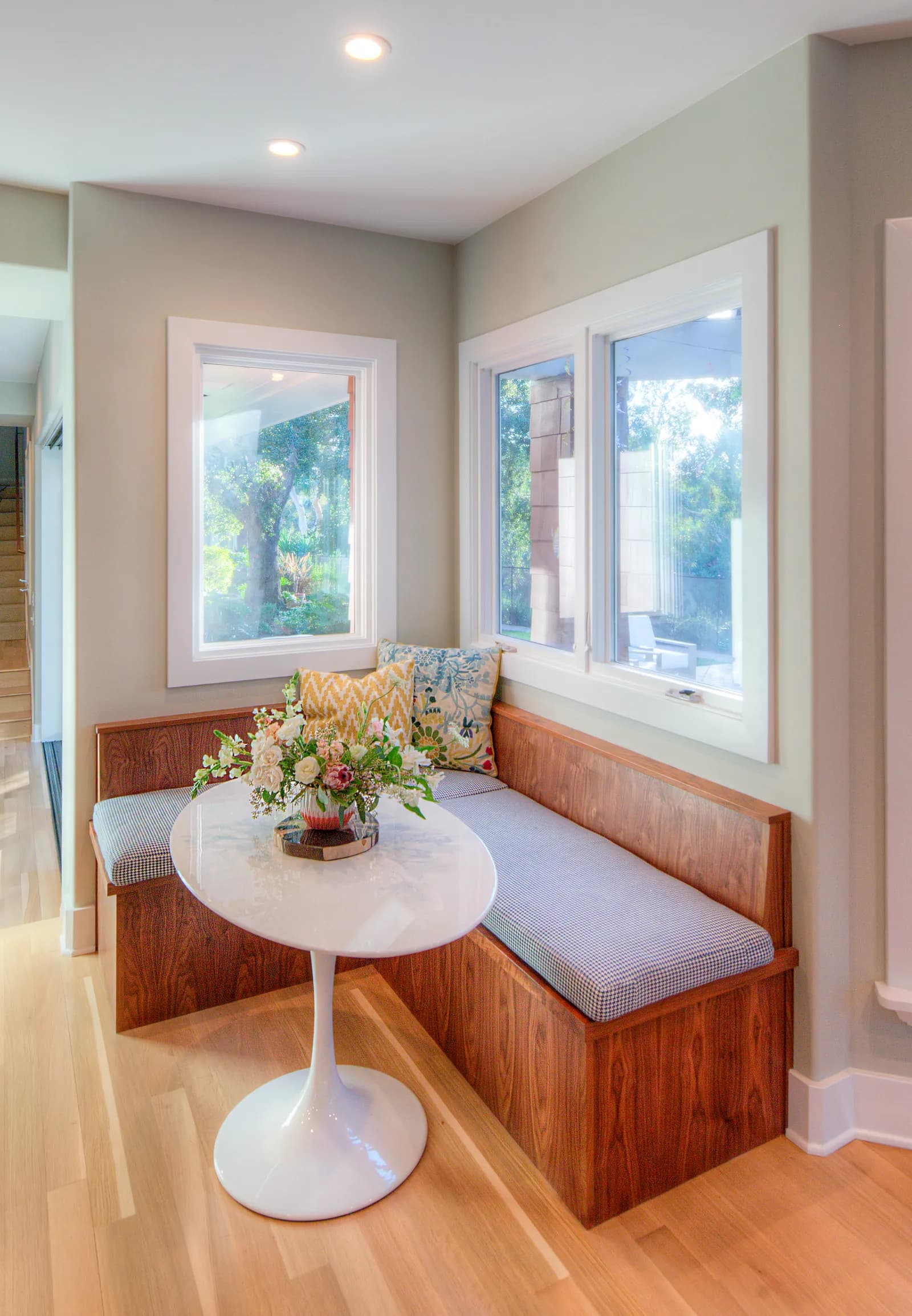 A corner breakfast nook with a built-in walnut banquette, patterned pillows, a white tulip table, and windows on two sides.