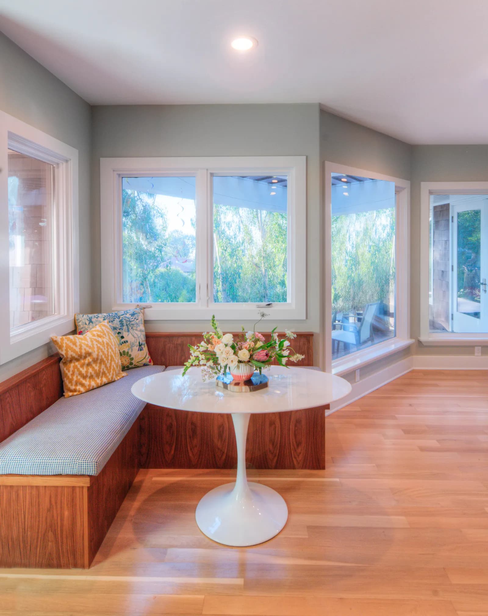 Breakfast nook with a built-in walnut banquette, patterned cushions, a white tulip table with flowers, and corner windows.