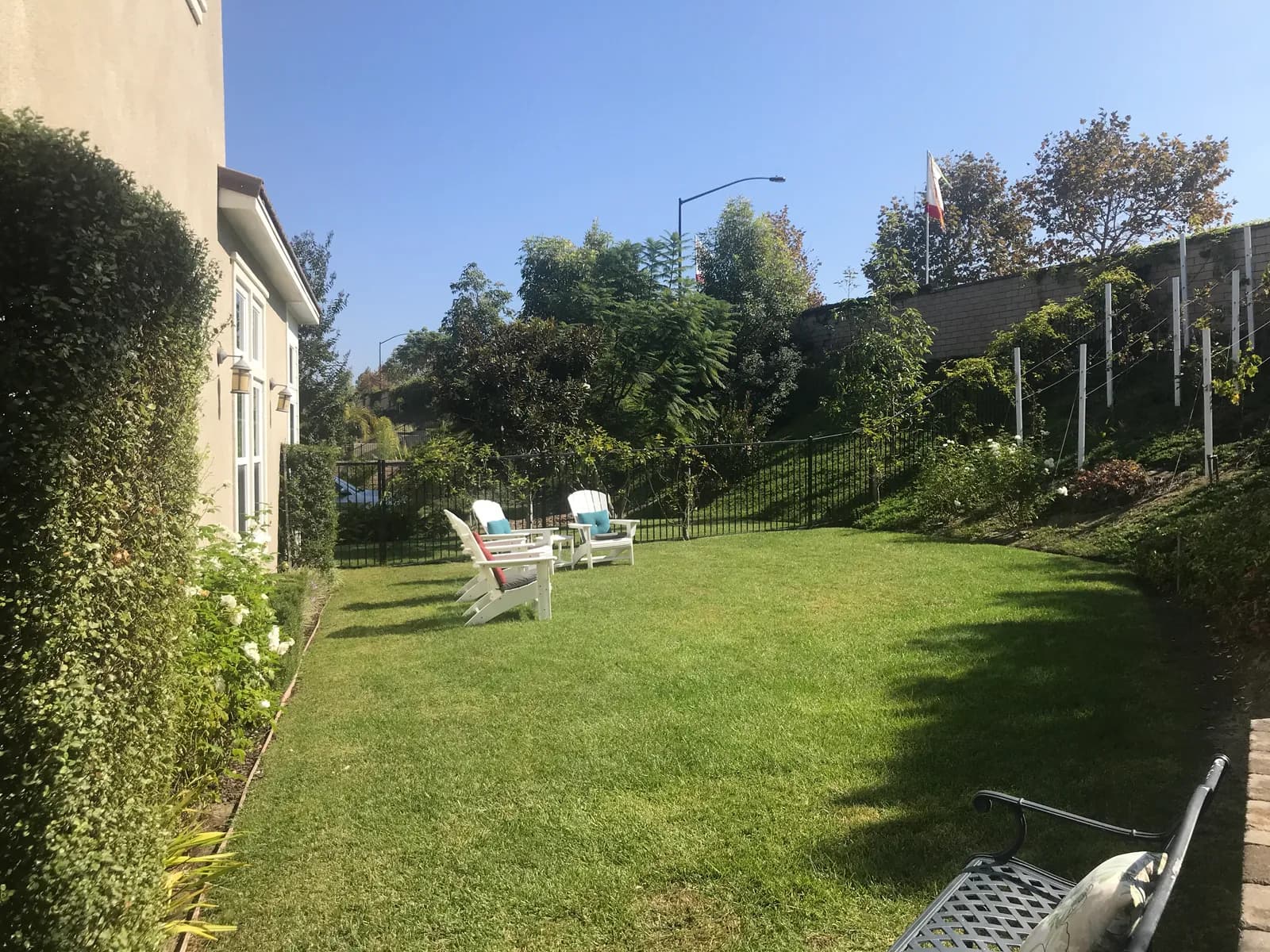 A grassy backyard with white Adirondack chairs, iron fencing, and a landscaped hillside behind.