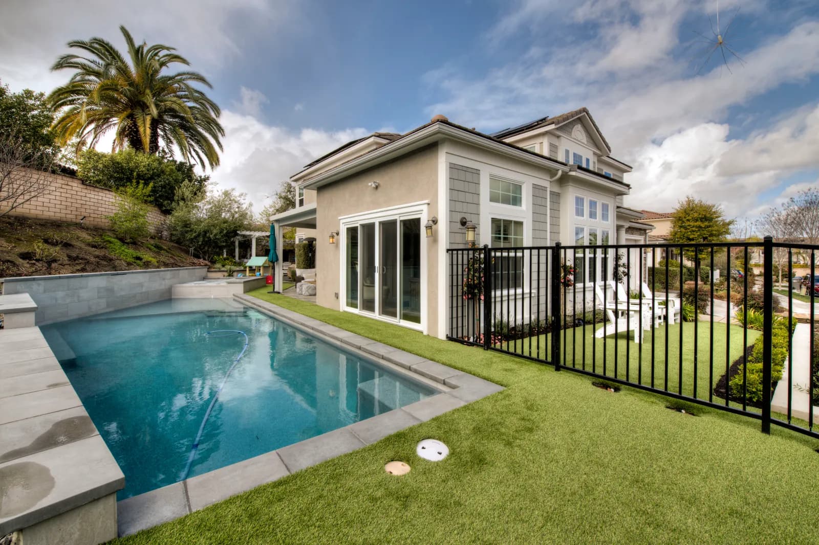 Backyard with a rectangular pool and spa, gray coping, artificial turf lawn, black metal pool fence, and a two-story home beyond.