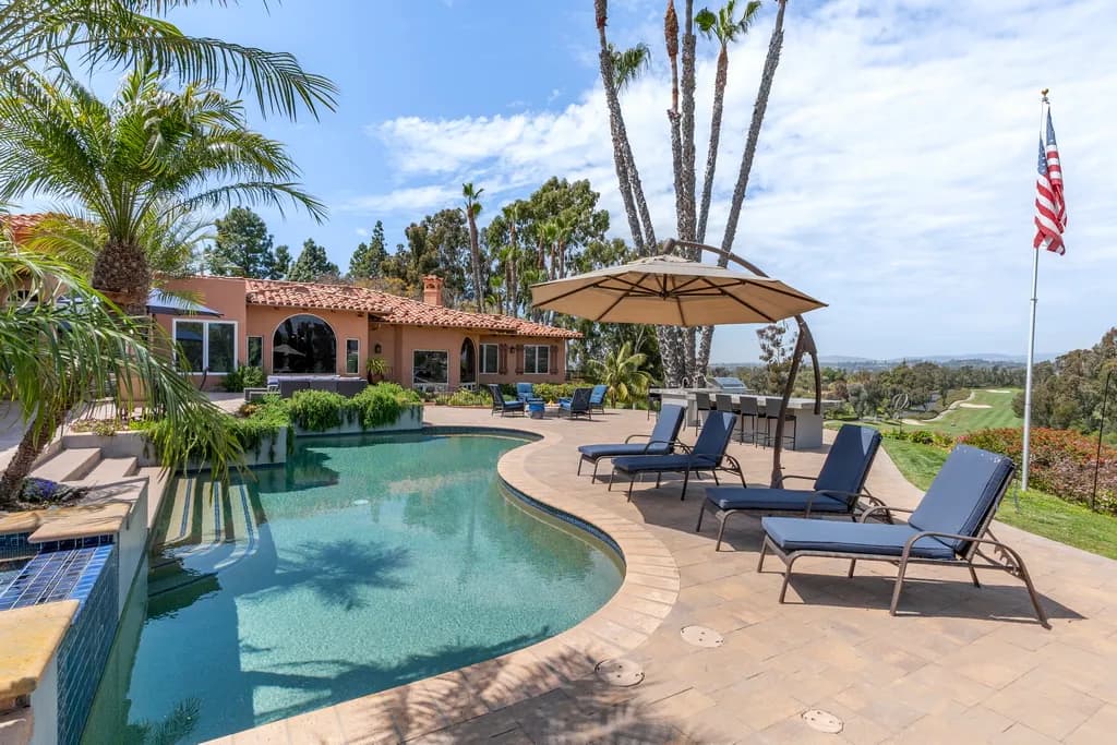 A curved backyard pool with chaise loungers, a cantilever umbrella, palm trees, and a valley view beyond.