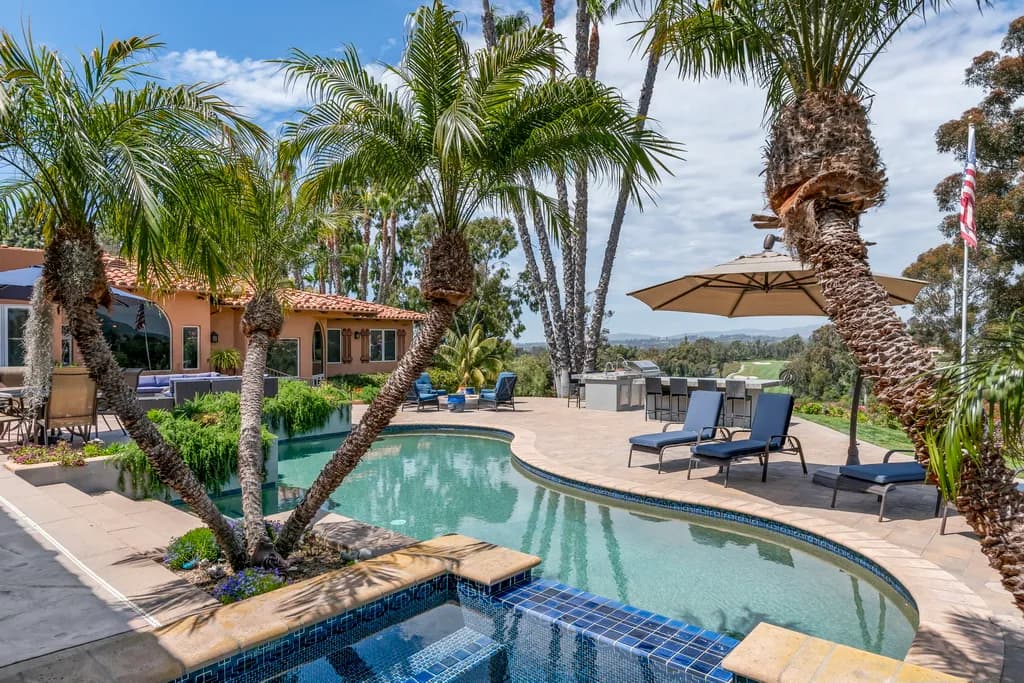 Backyard pool and spa surrounded by palm trees, lounge chairs, an umbrella, and an outdoor kitchen with hillside views.