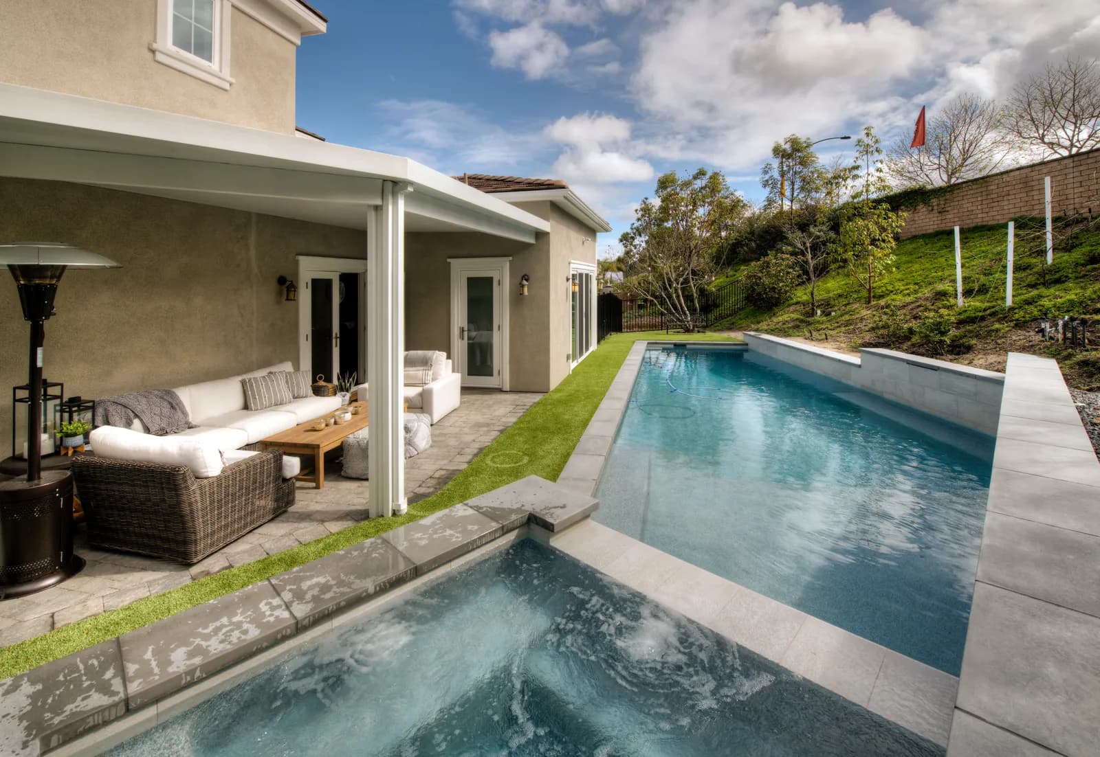 A backyard with a raised spa spilling toward a long rectangular pool beside a covered patio with wicker seating.